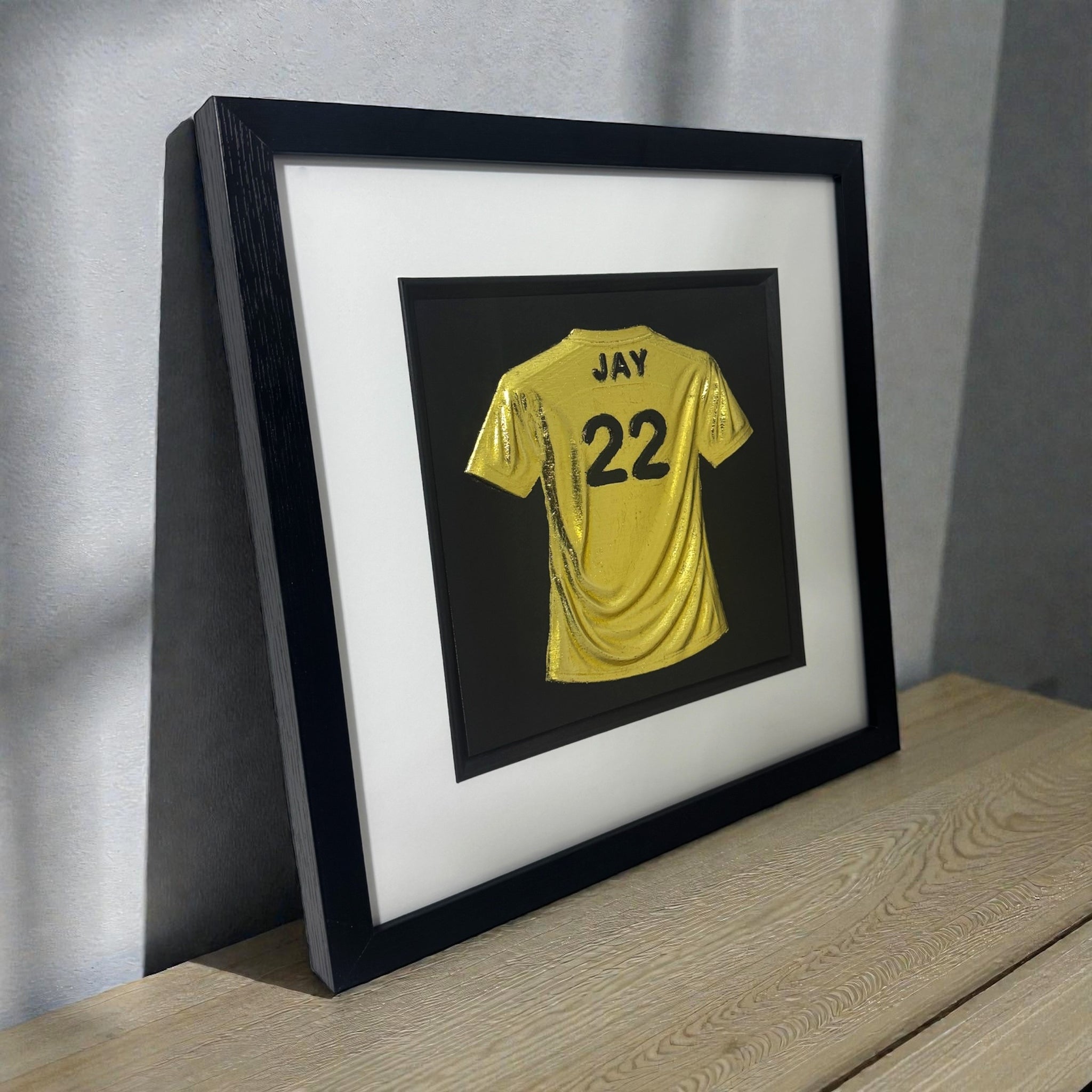 Framed personalised yellow football shirt 3D wall art with name “JAY” and number “22”, in a black frame with white mount, standing on a wooden surface.