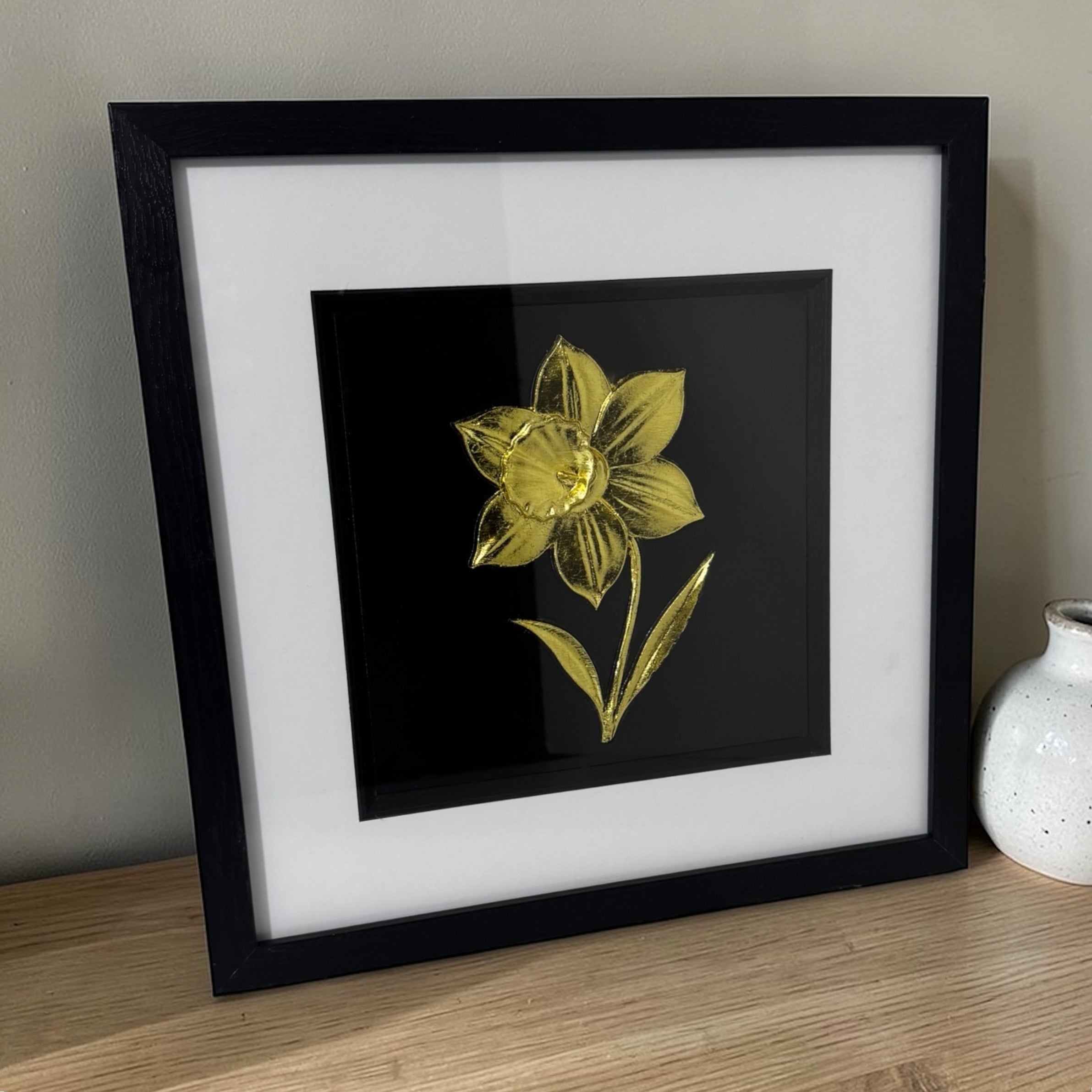 Angled view of a gold daffodil relief in a black box frame on a wooden sideboard against a light wall.