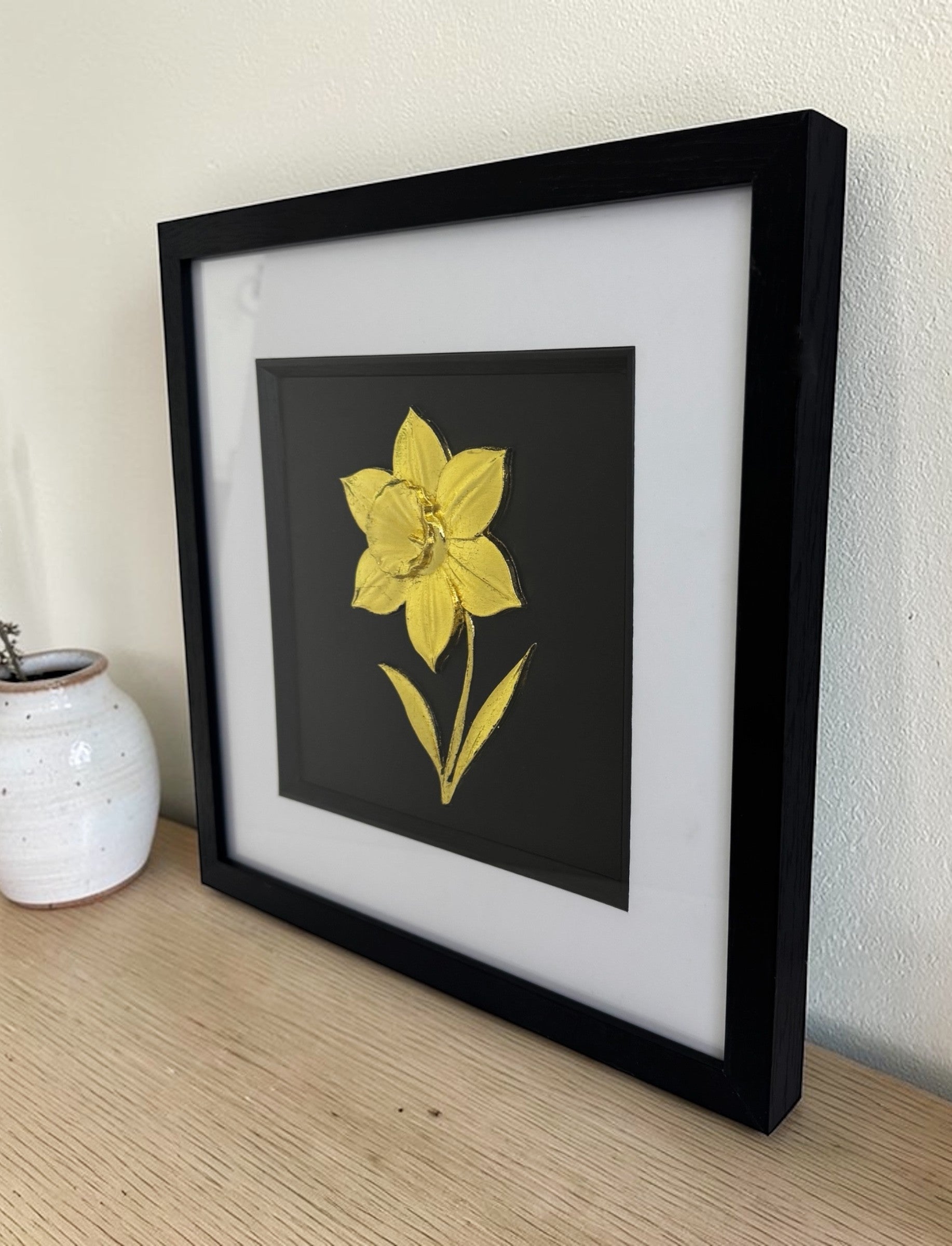 Gold daffodil relief in a black box frame with glass front on a wooden sideboard beside a white ceramic vase.