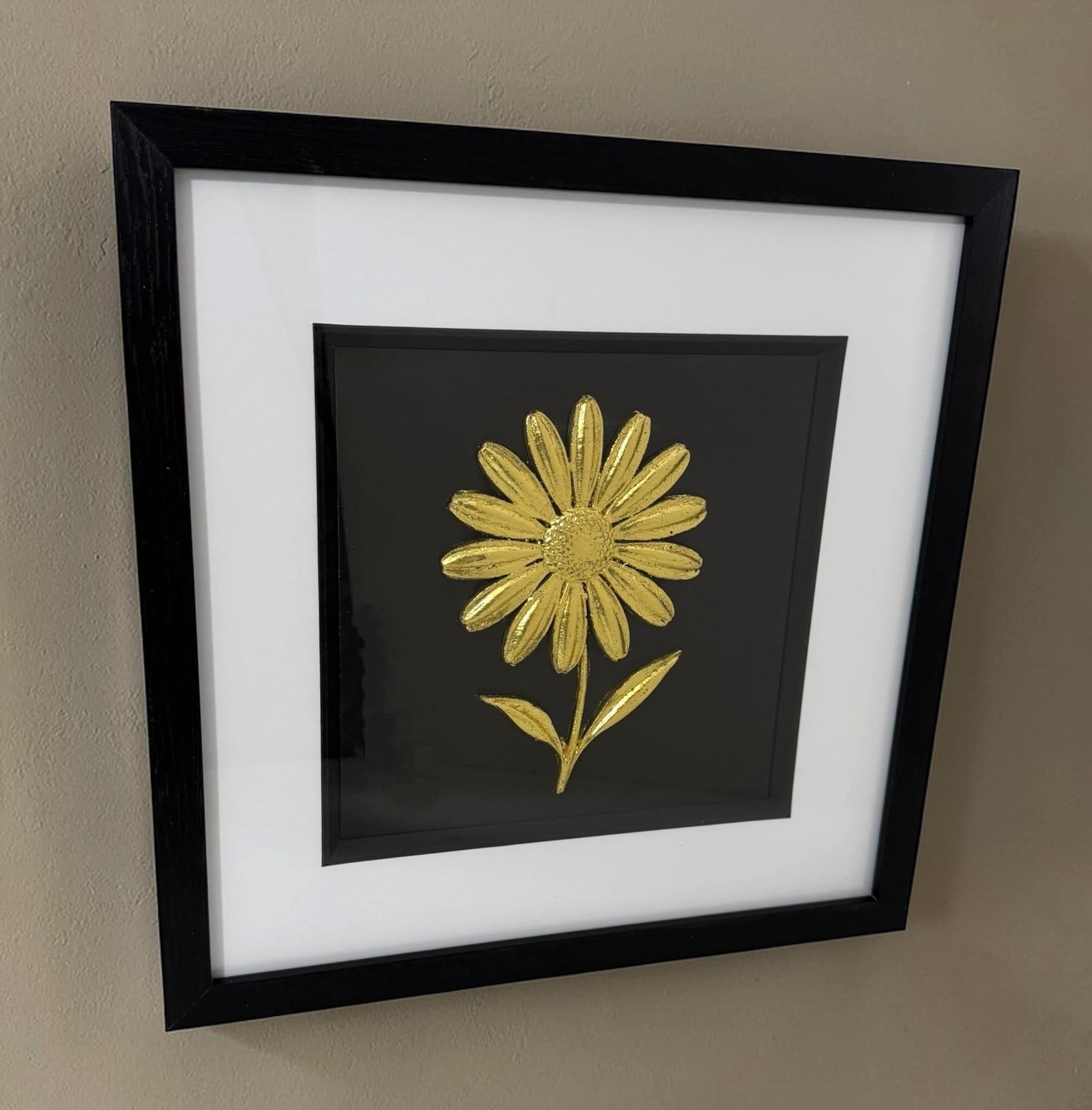 Gold daisy raised relief in a black frame with white mount, black inner surround, and glass front on a neutral wall.