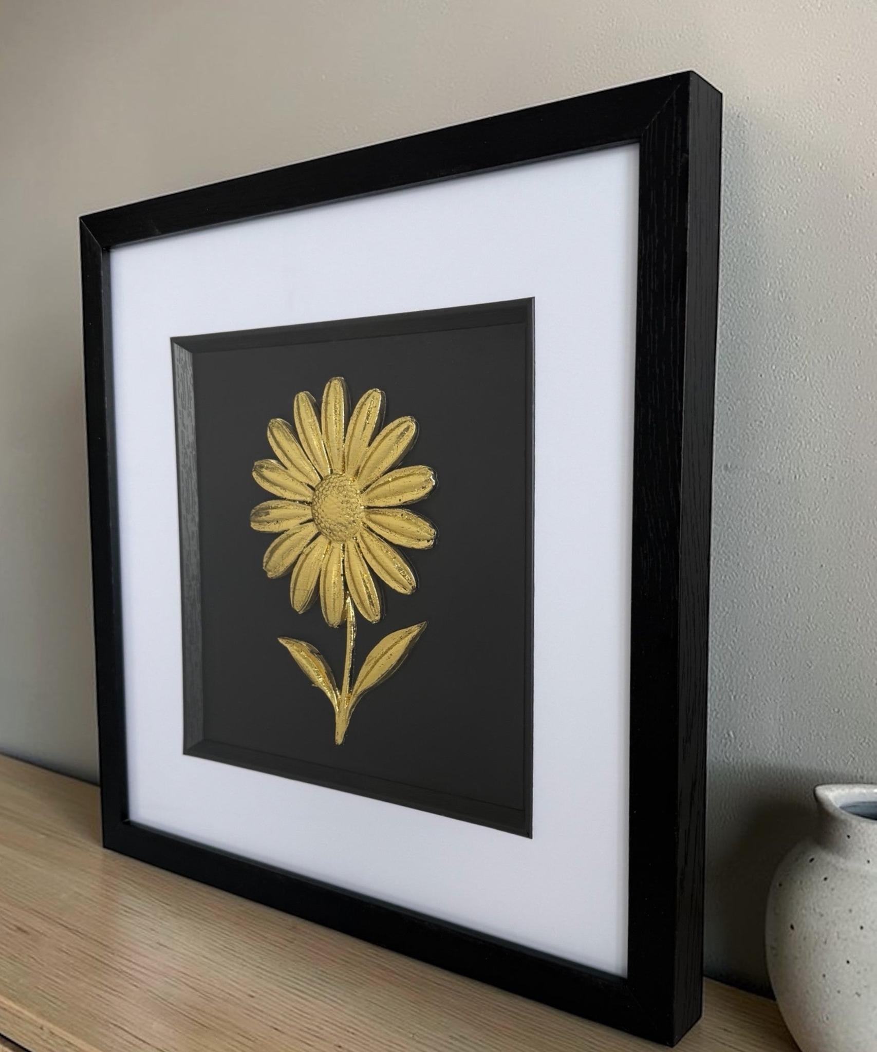 Angled view of gold daisy raised relief wall decor in a black frame on a wooden sideboard beside a ceramic vase.