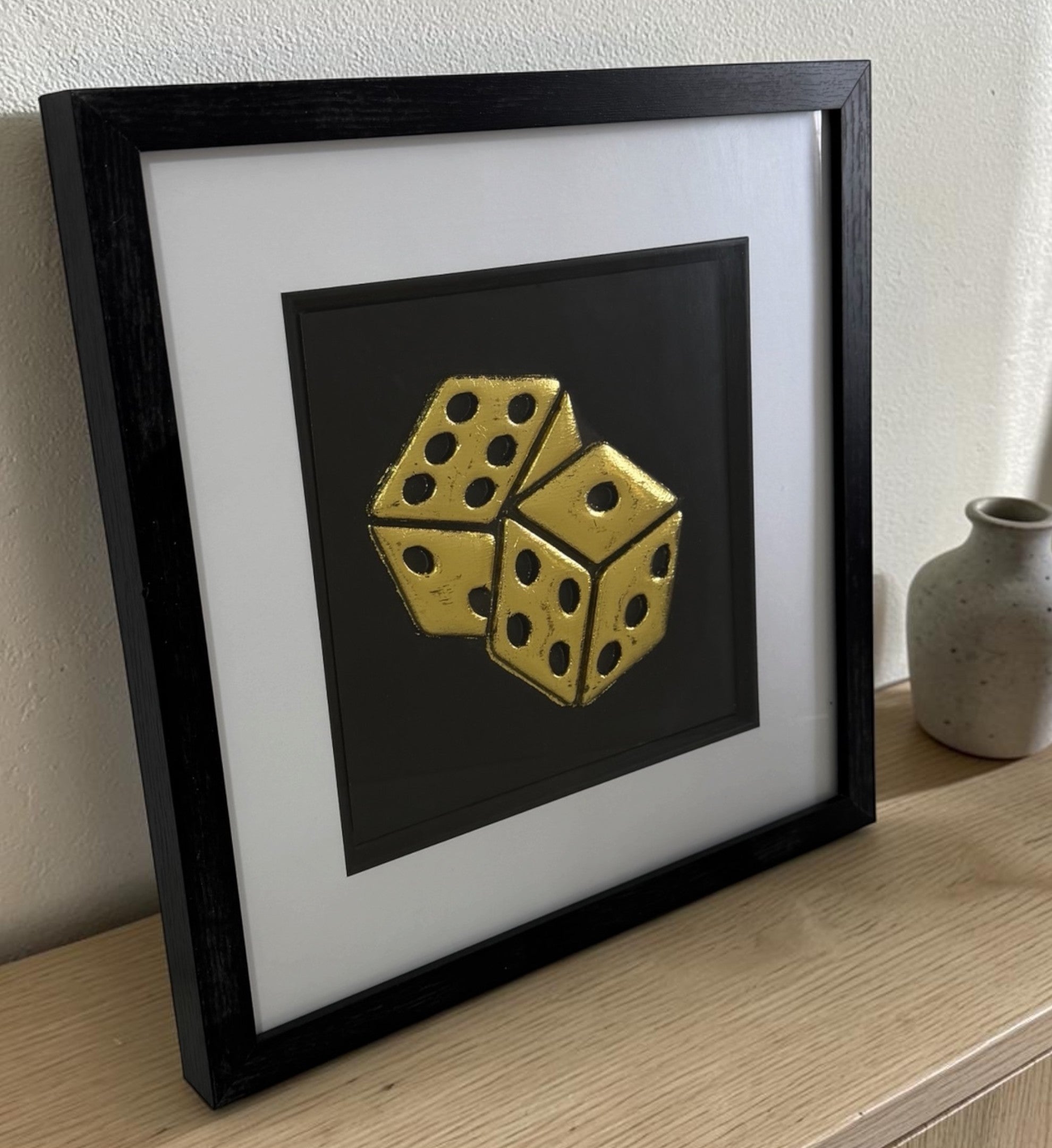 Gold leaf dice wall art in a black frame with white mount, styled on a wooden shelf with neutral decor for games room styling.