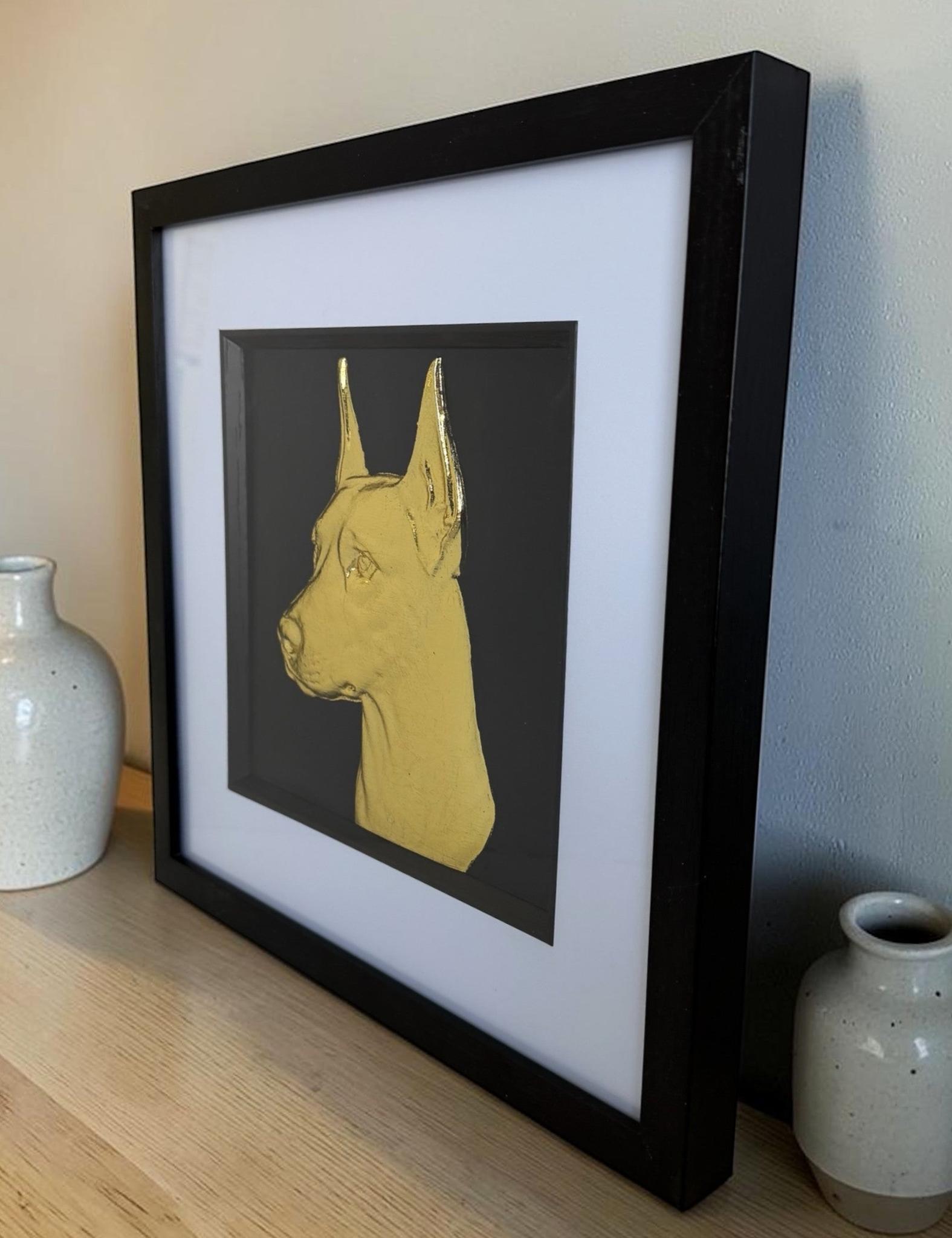 Gold doberman relief portrait in a black box frame with white mount on a light wood surface beside ceramic vases.