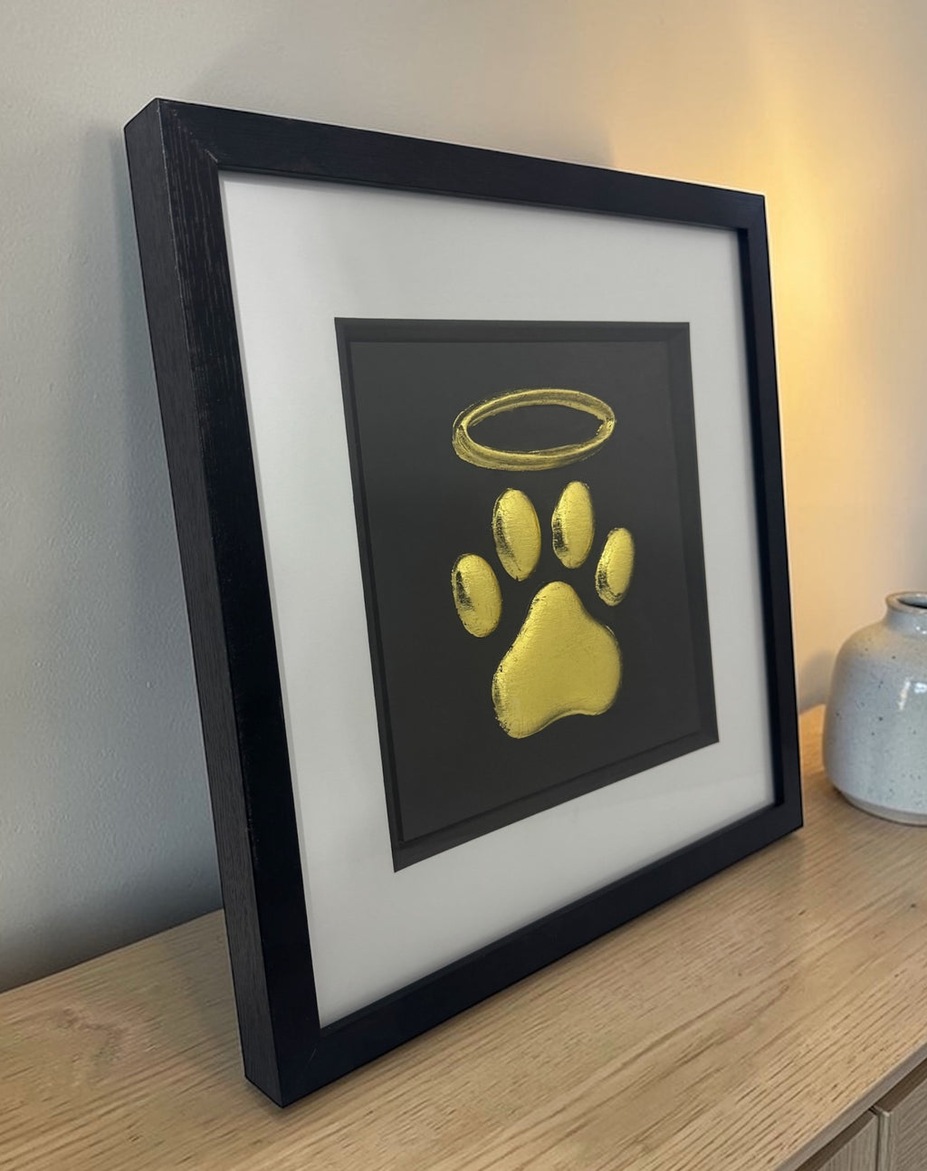 dog paw and halo memorial 3d wall art in frame gold leaf with black background and white mount, on a desk