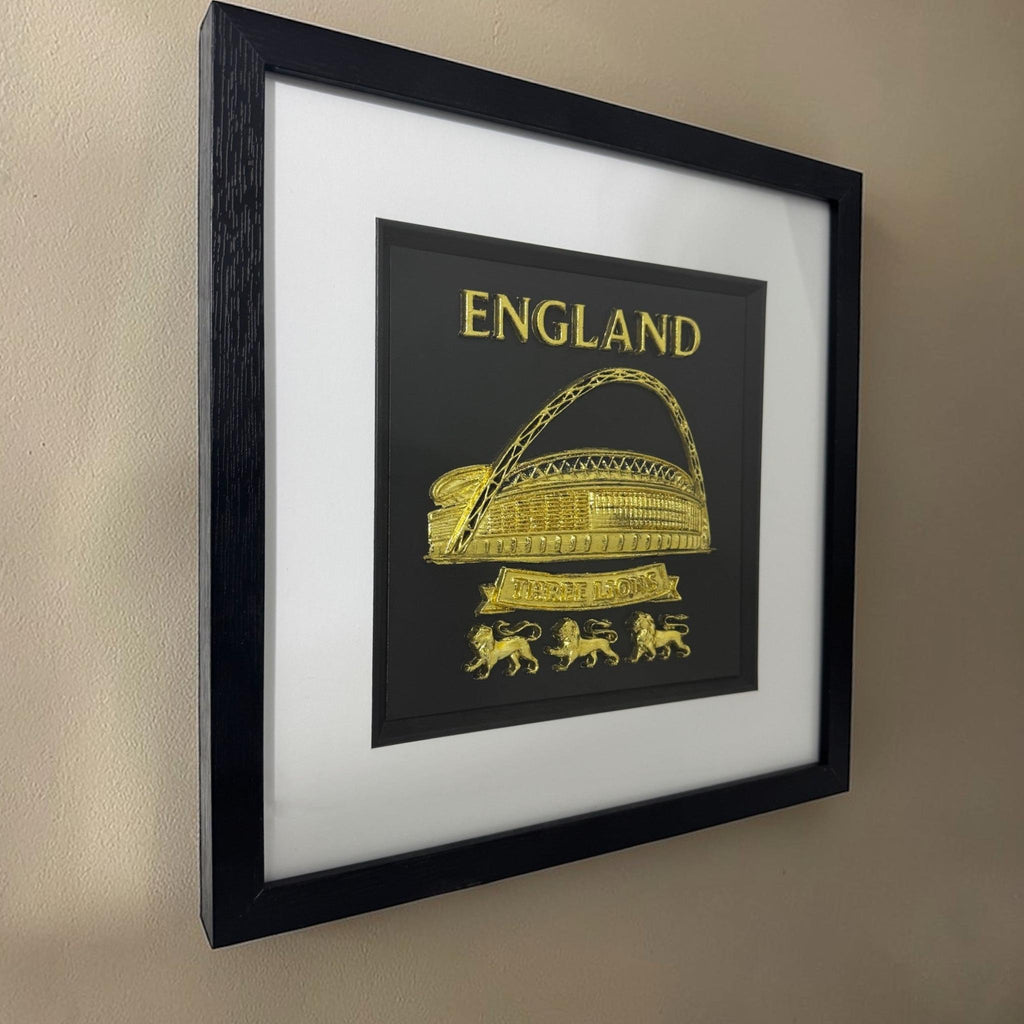 Gold England Wembley Stadium 3D relief artwork in black square frame with white mount, angled view on wall showing frame depth and glass reflections