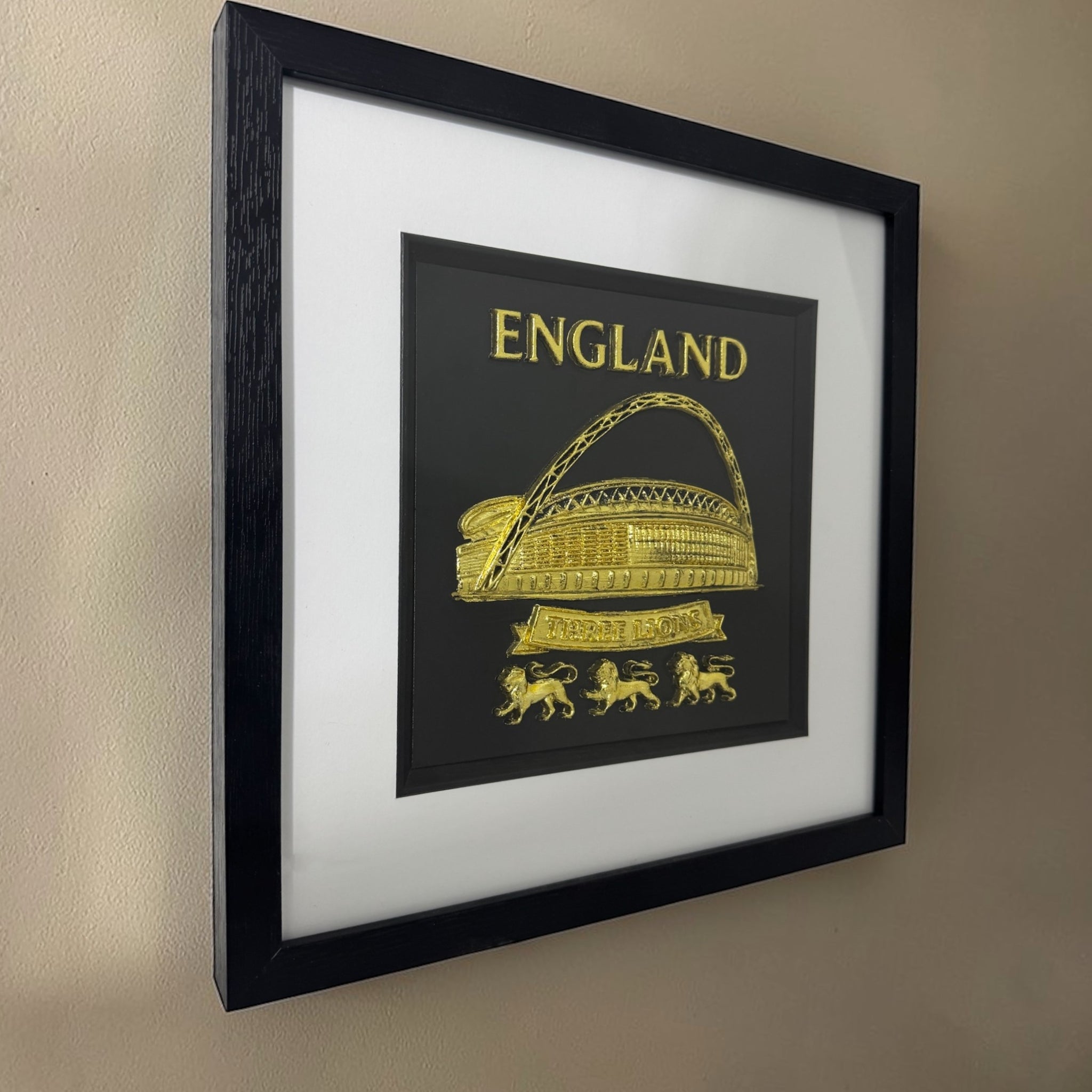 Gold England Wembley Stadium 3D relief artwork in black square frame with white mount, angled view on wall showing frame depth and glass reflections