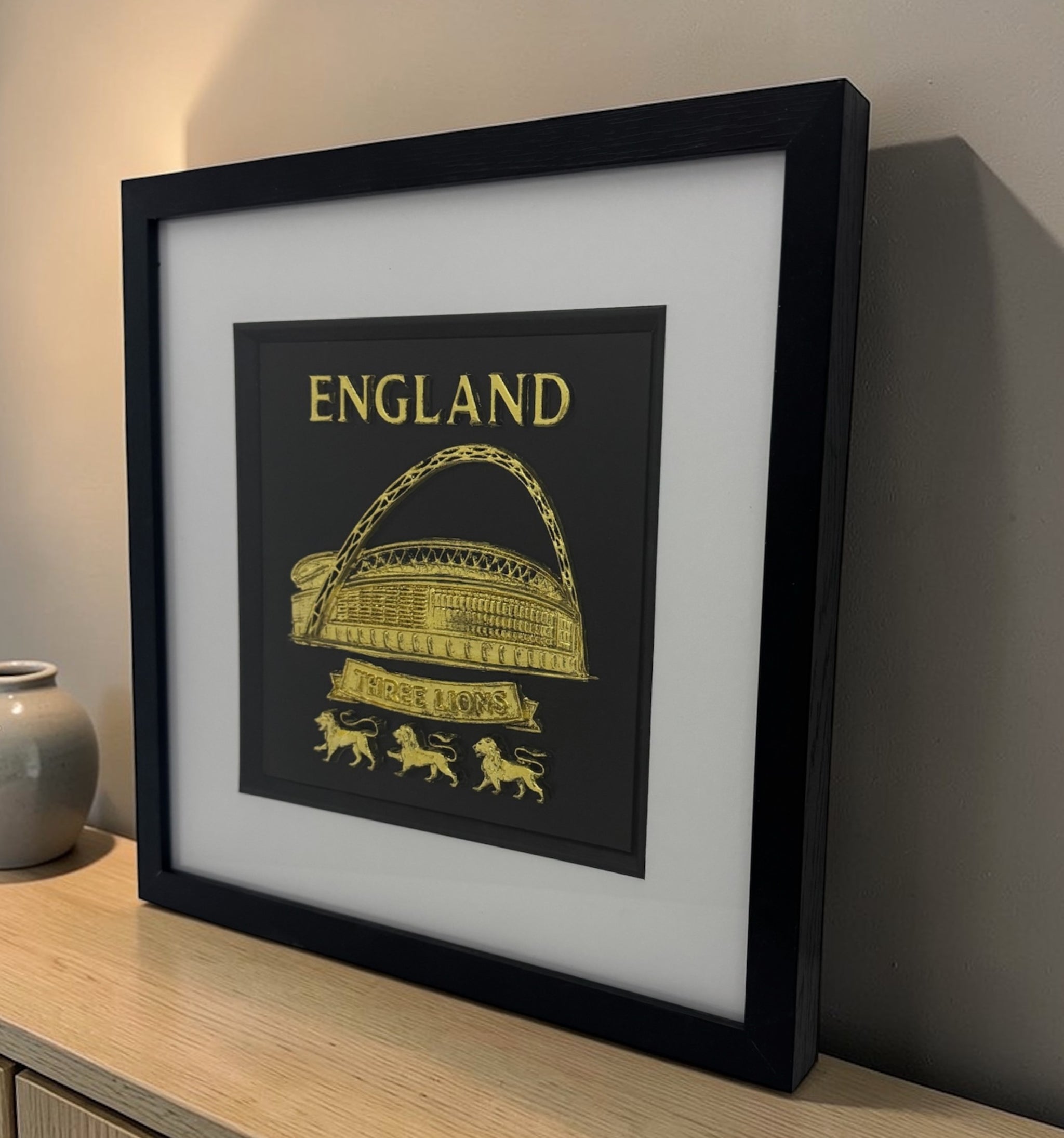 Gold England Wembley Stadium 3D relief artwork in black square frame with white mount, angled view on desk