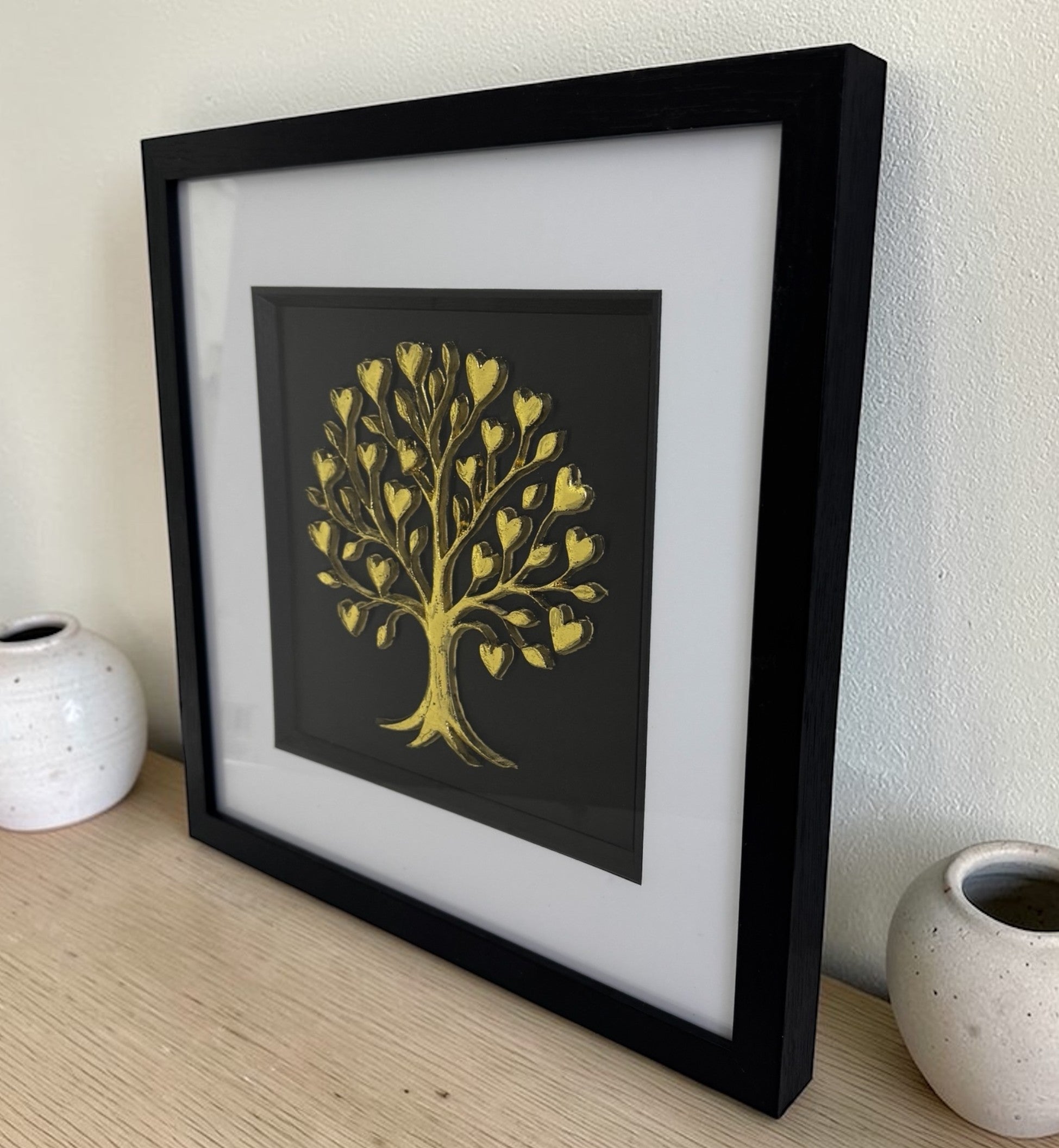Gold tree of life relief styled on a wooden sideboard in a black box frame beside ceramic vases.