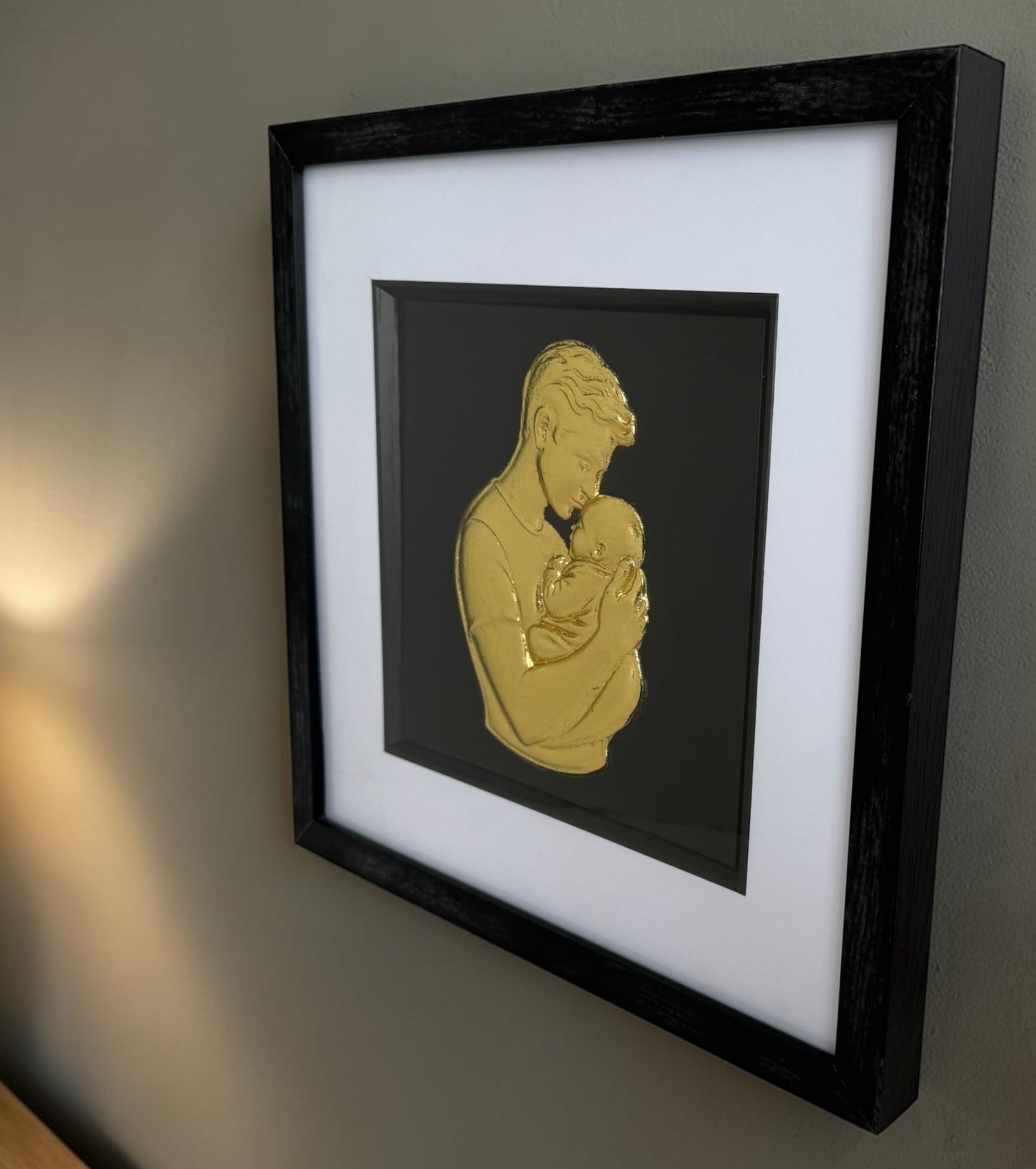 Angled wall view of gold father and baby raised relief artwork showing black box frame depth.