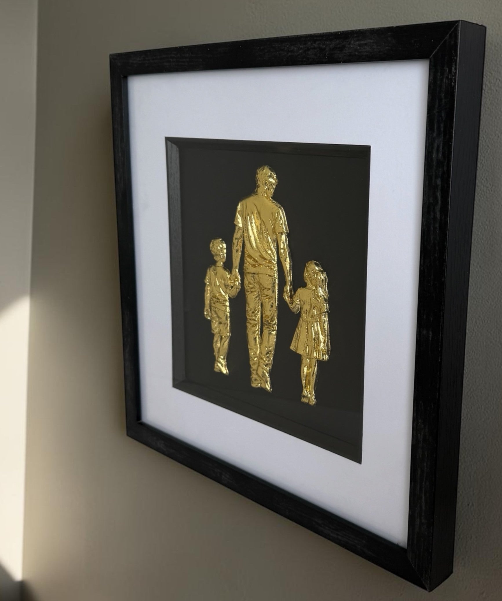 Angled wall view of gold father and children raised relief artwork showing square black frame depth.