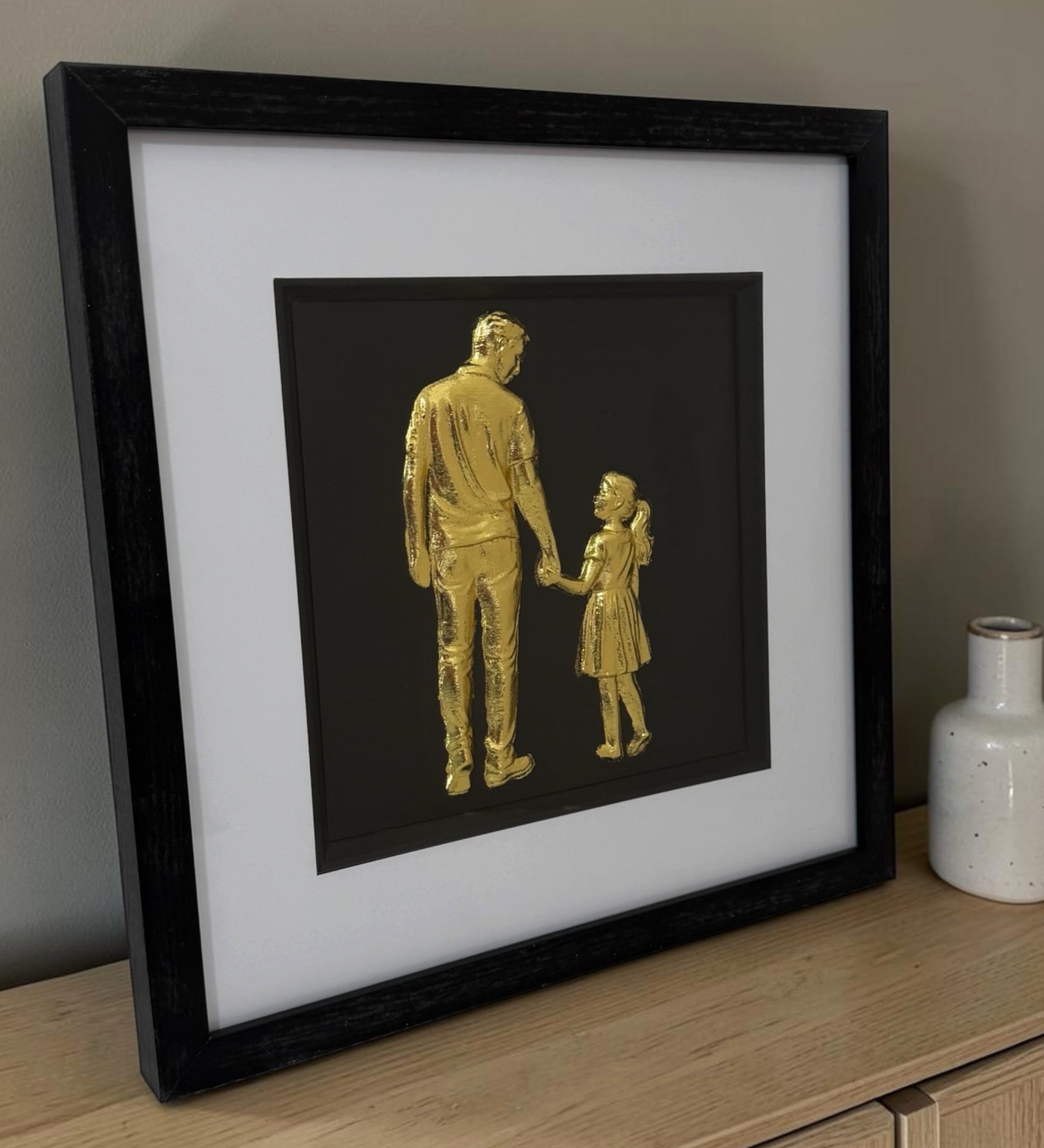 Angled close-up of gold father and daughter framed wall art on a wooden surface, featuring a sentimental parent and child design with black frame and white mount.