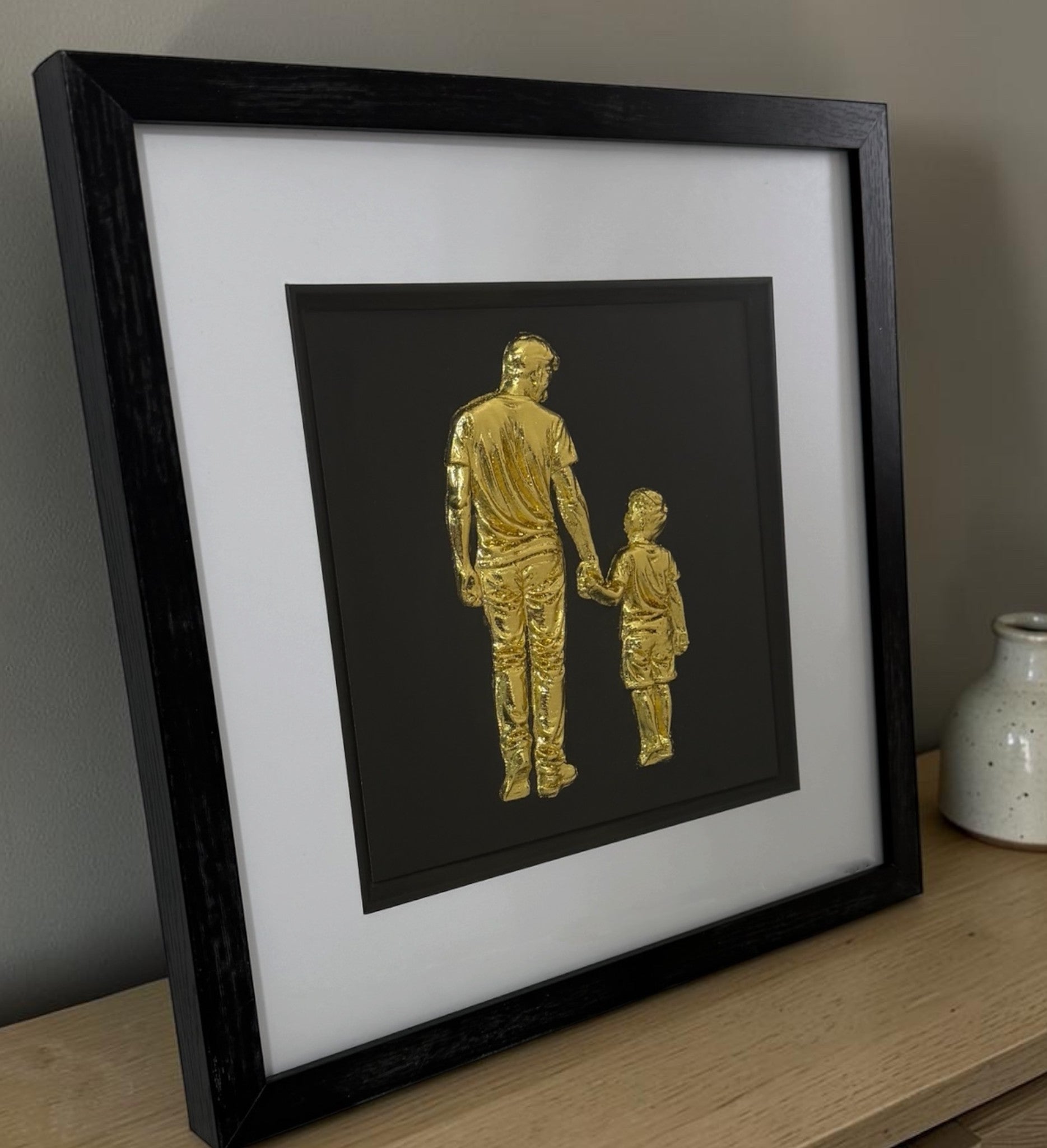 Gold father and son framed 3D artwork on a sideboard showing black box frame depth and raised detail.