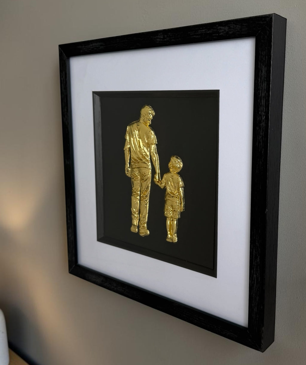 Angled wall view of gold father and son raised relief artwork showing square black frame depth.