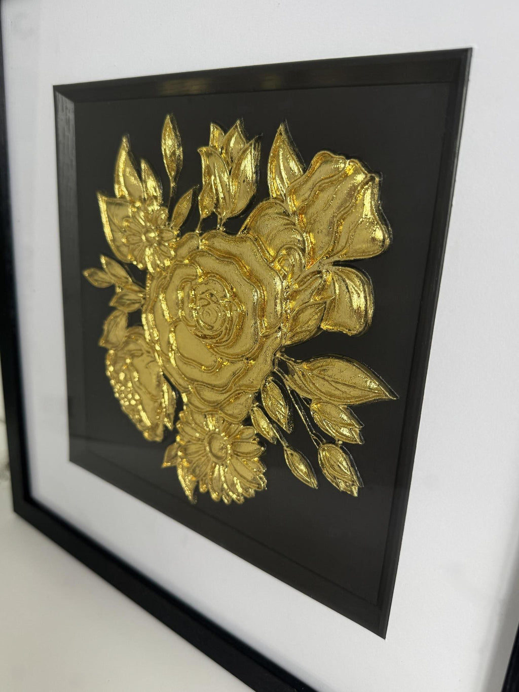 Close-up angled view of raised gold-finish floral bouquet relief showing layered petals and leaf details on a black centre.