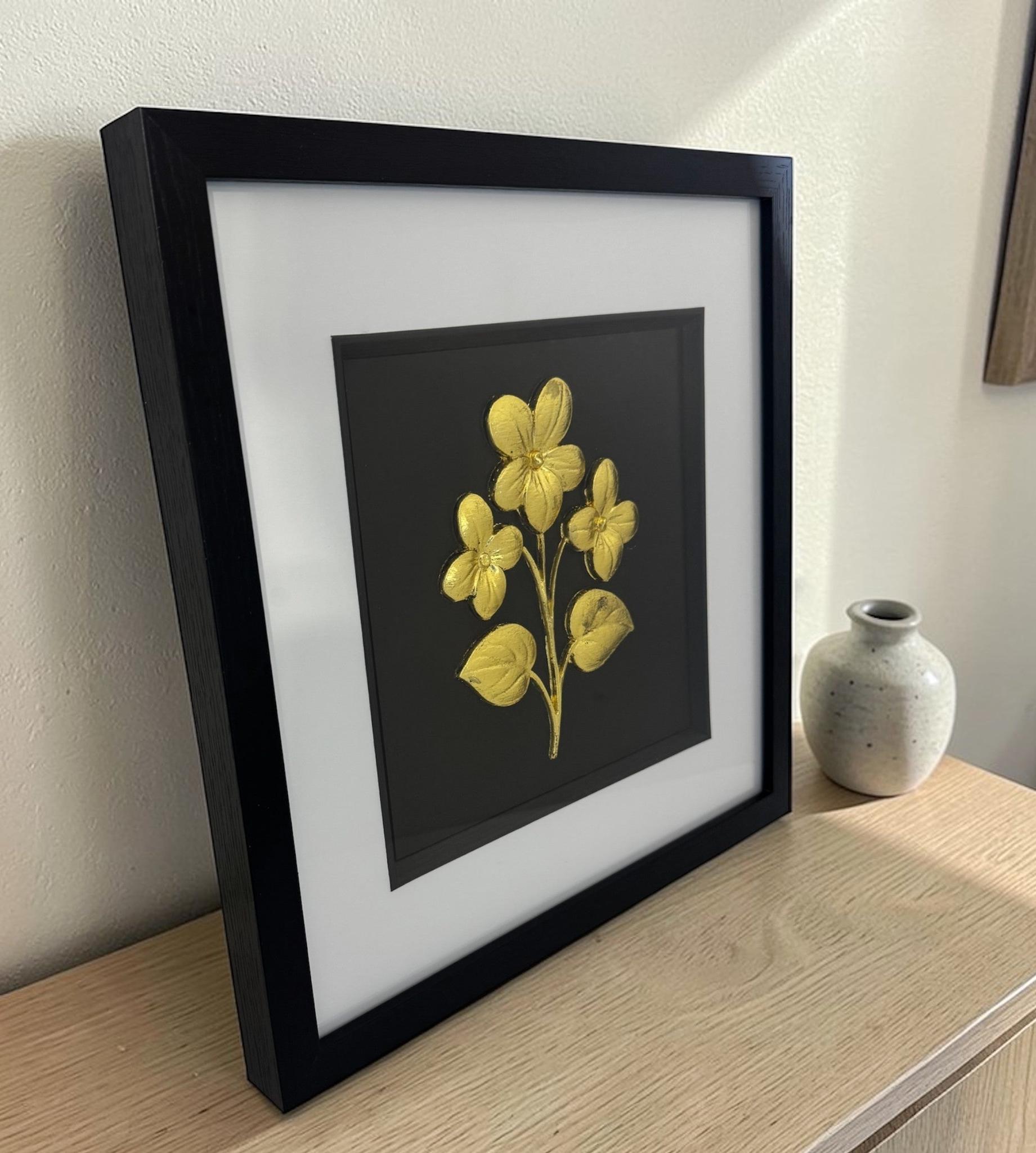 Angled view of the gold framed floral relief with glass front on a light wood sideboard beside a neutral ceramic vase