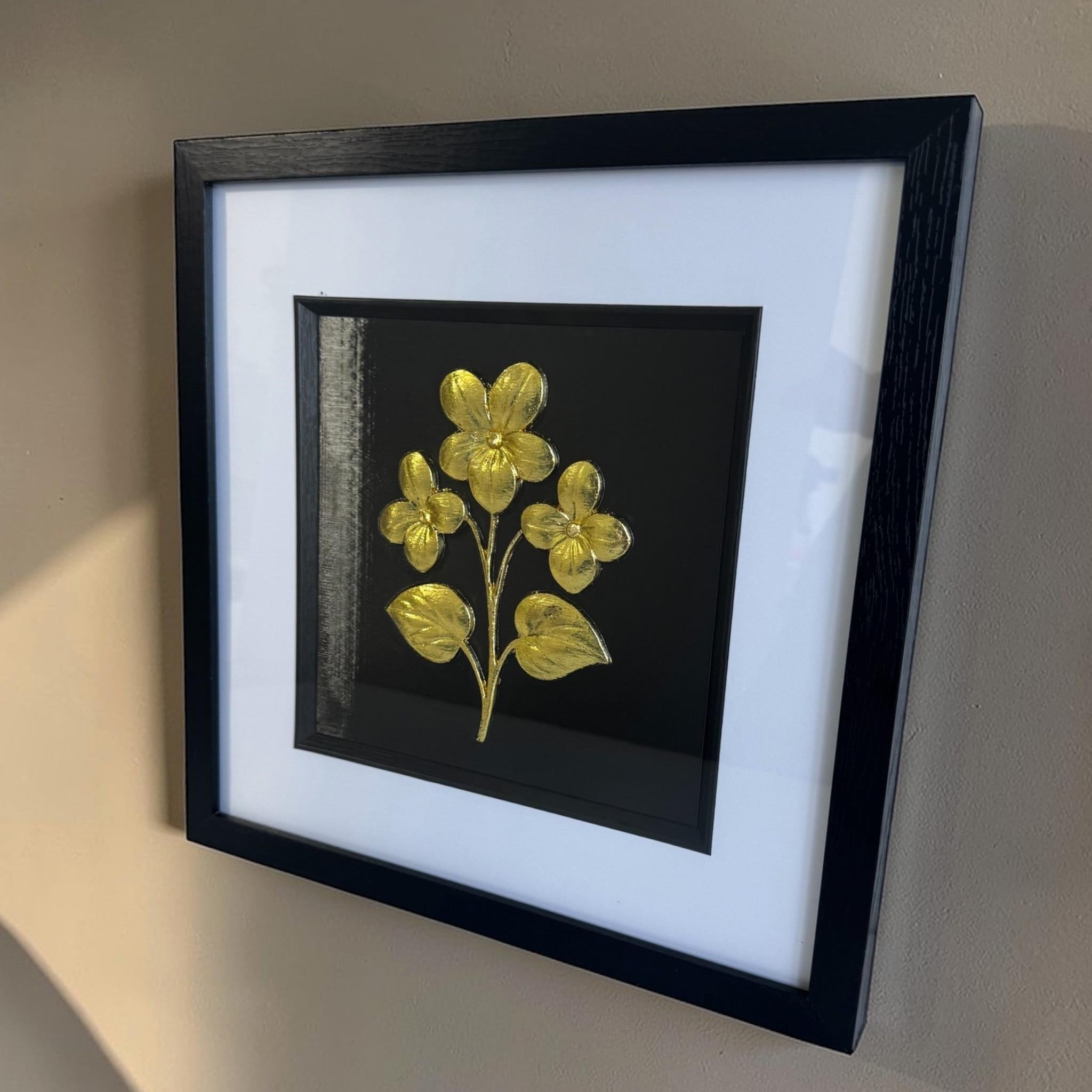 Gold framed floral relief hanging on a beige wall with a wide white mount and black inner surround