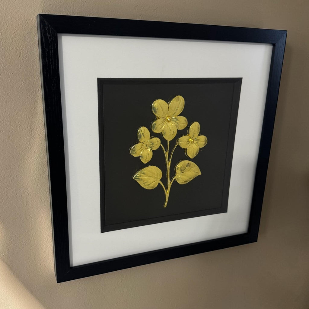 Angled gold flower wall decor in a black frame with a white mount and black inner surround on a beige wall