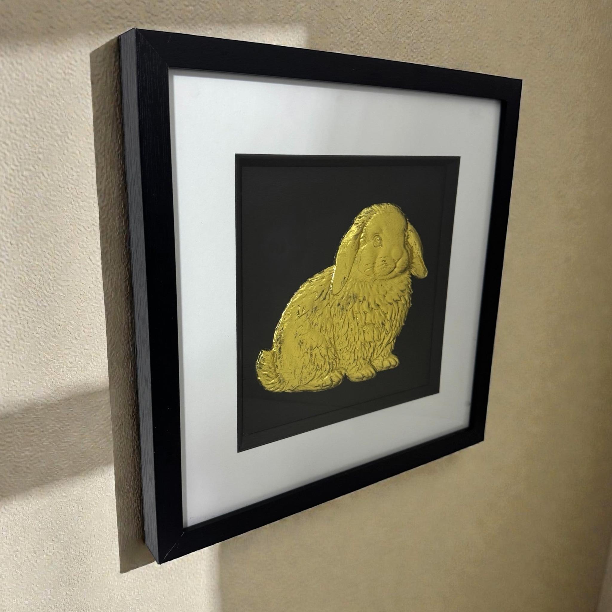 Side view of framed Mini Lop rabbit 3D wall art in gold leaf, in a black frame with white mount, hung on a beige wall.