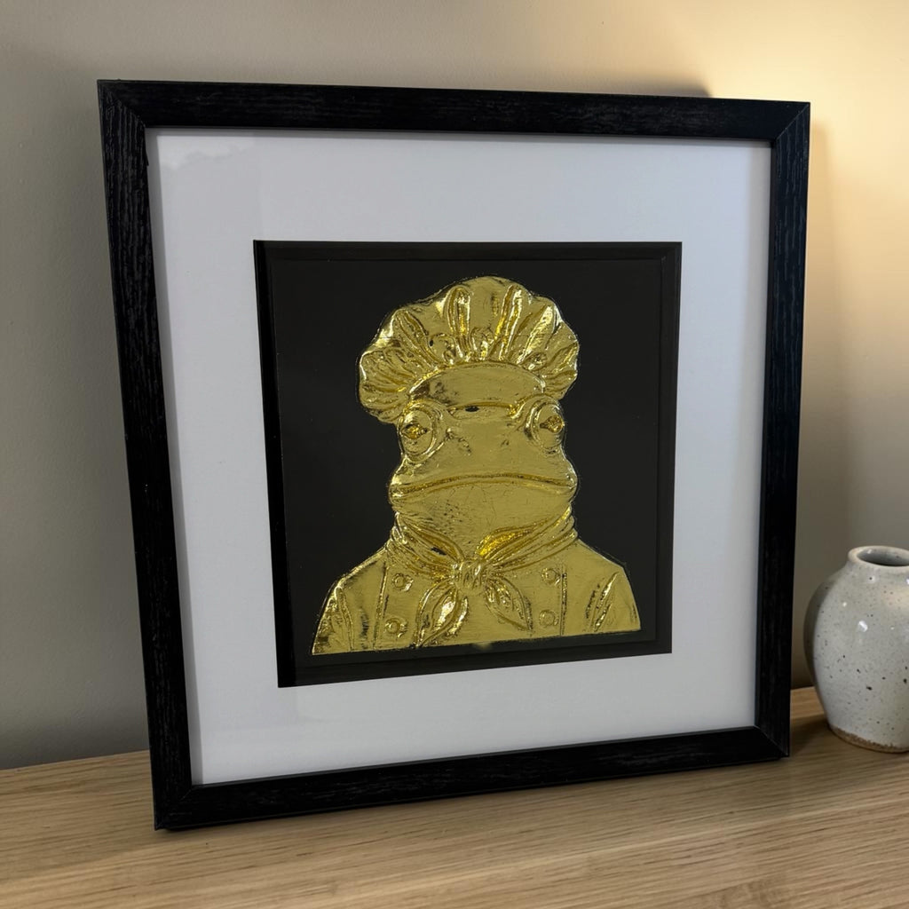 Gold frog chef framed artwork on a wooden surface with warm side lighting and visible black box frame