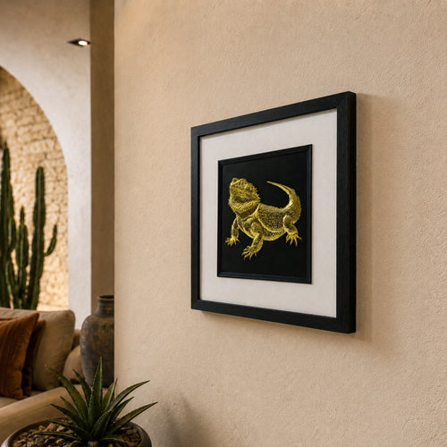 Angled lifestyle view of gold bearded dragon 3D wall art in a black frame with white mount, hanging on a warm neutral wall.