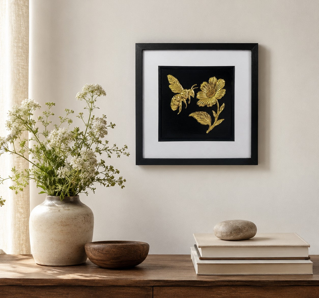 Gold bee and flower framed wall art displayed on a neutral wall above a wooden console with wildflowers, showing botanical gold leaf artwork in a 33cm square black frame.