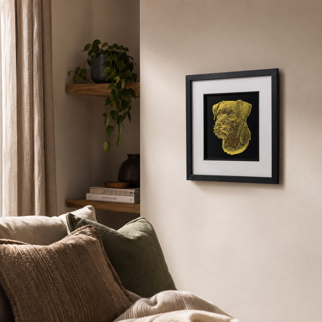 Angled lifestyle view of gold Border Terrier framed wall art hung on a neutral wall beside soft cushions, warm natural light, greenery and wooden shelving.