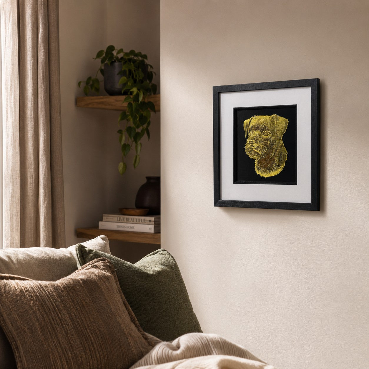 Angled lifestyle view of gold Border Terrier framed wall art hung on a neutral wall beside soft cushions, warm natural light, greenery and wooden shelving.