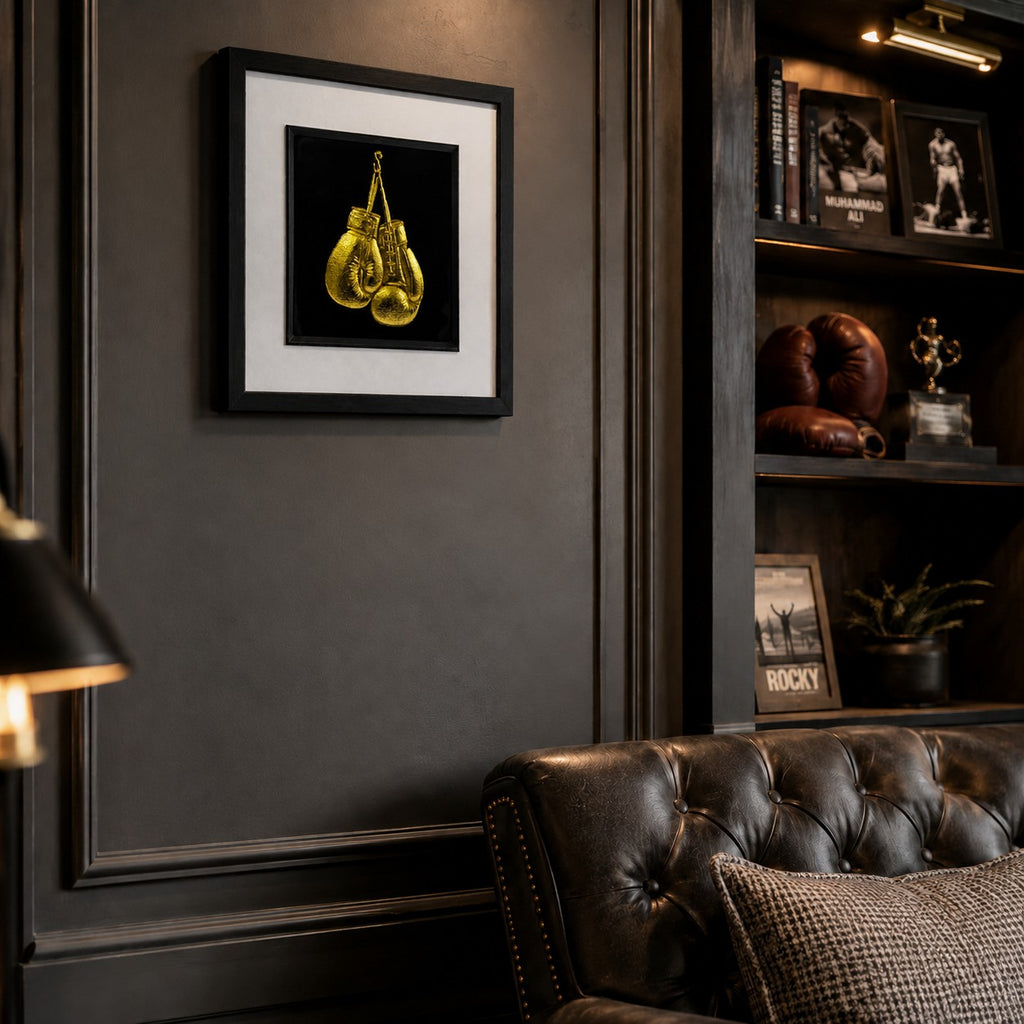 Angled lifestyle view of framed gold boxing gloves wall art mounted on a dark study wall, styled with boxing memorabilia, leather seating and warm luxury decor.