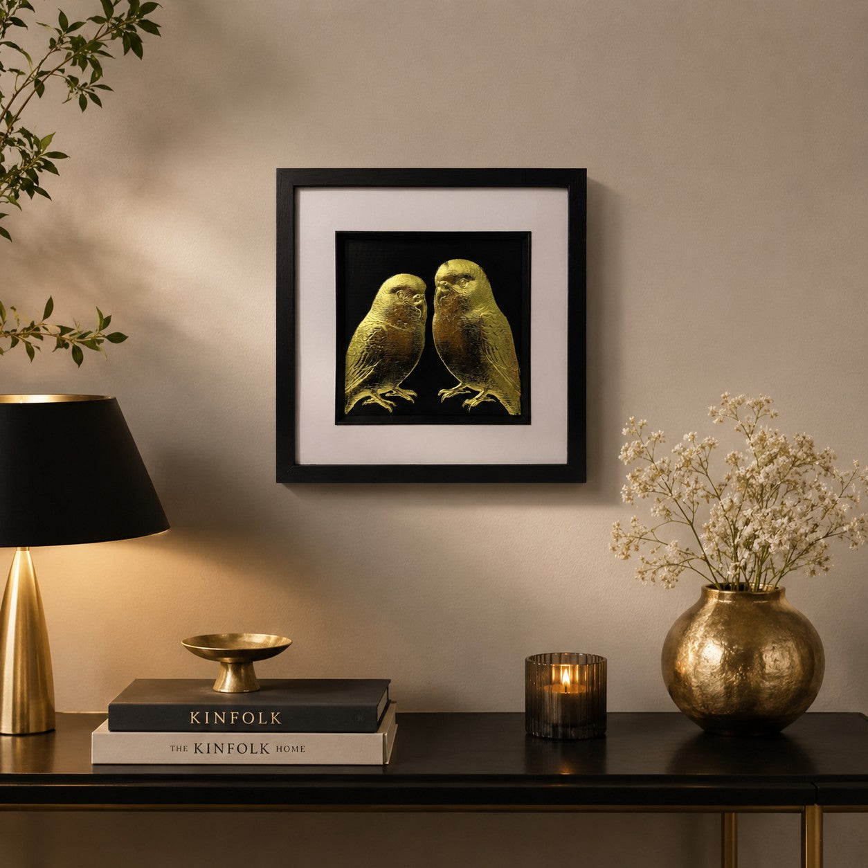 Gold budgie bird wall art in a black square frame styled above a dark console with warm lighting, gold accents, flowers and neutral home decor.