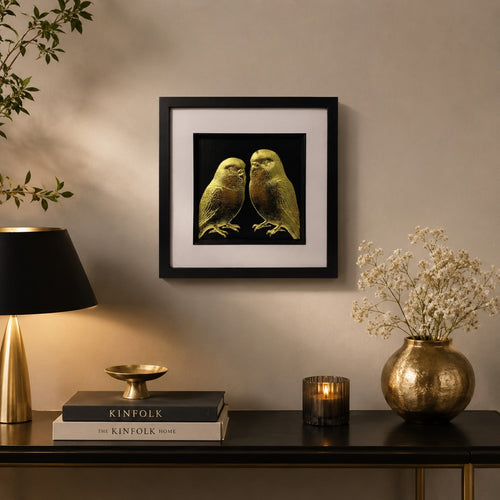 Gold budgie bird wall art in a black square frame styled above a dark console with warm lighting, gold accents, flowers and neutral home decor.