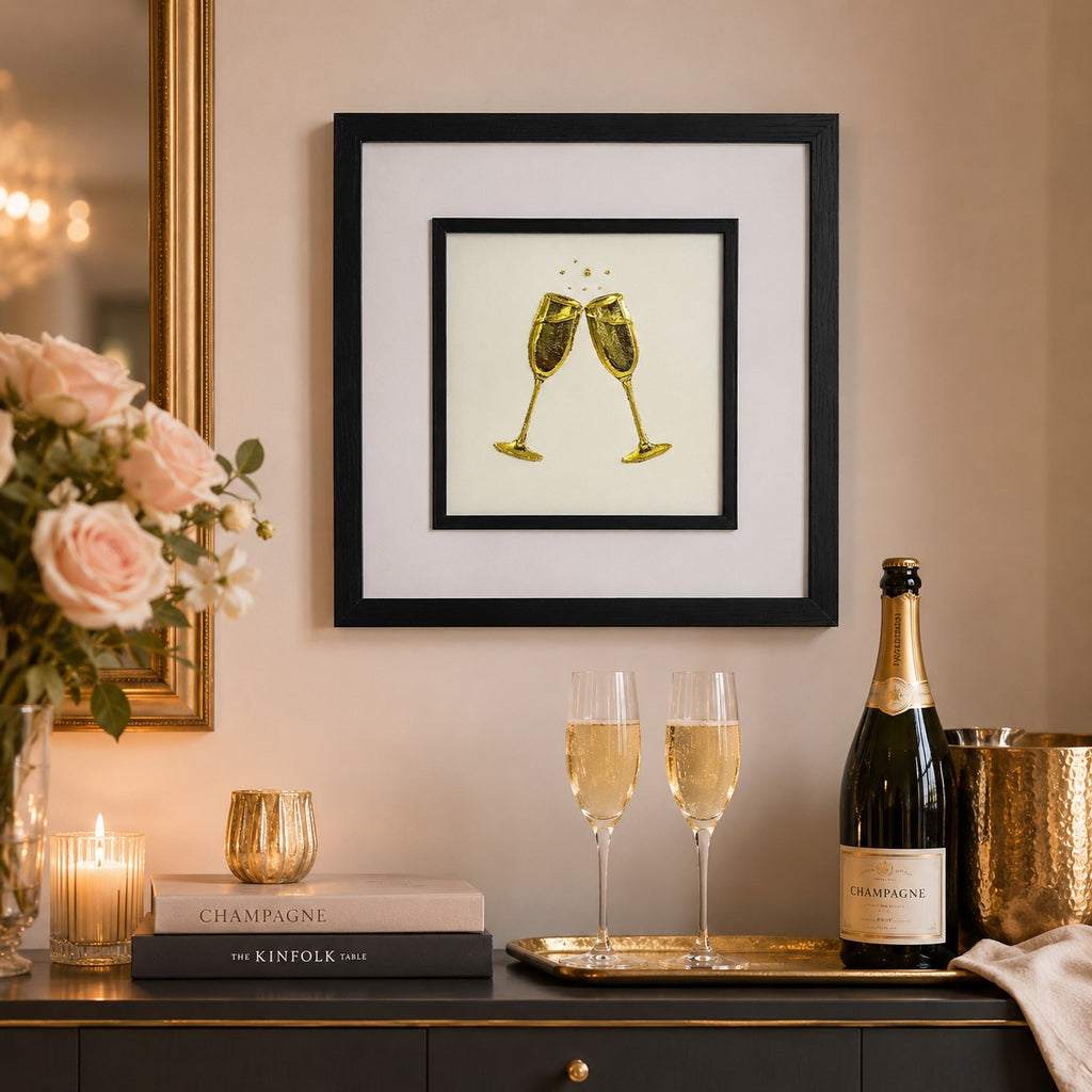 Gold champagne glasses wall art in a black square frame styled above a luxury celebration console with champagne flutes, roses, candlelight and warm gold decor.