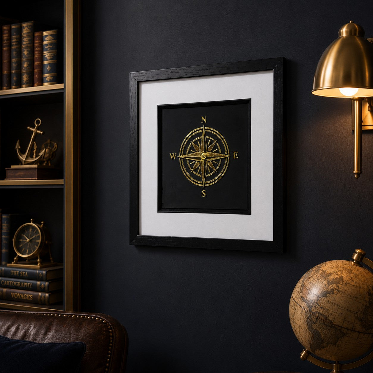Angled wall view of gold compass framed wall art in a moody nautical study with brass lamp, vintage globe, leather chair and travel-inspired decor.