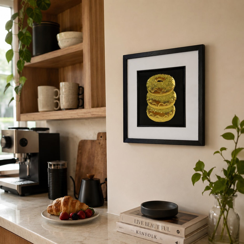 Gold 3D doughnut stack artwork in black frame styled on a kitchen wall with coffee cup, wood accents and greenery.
