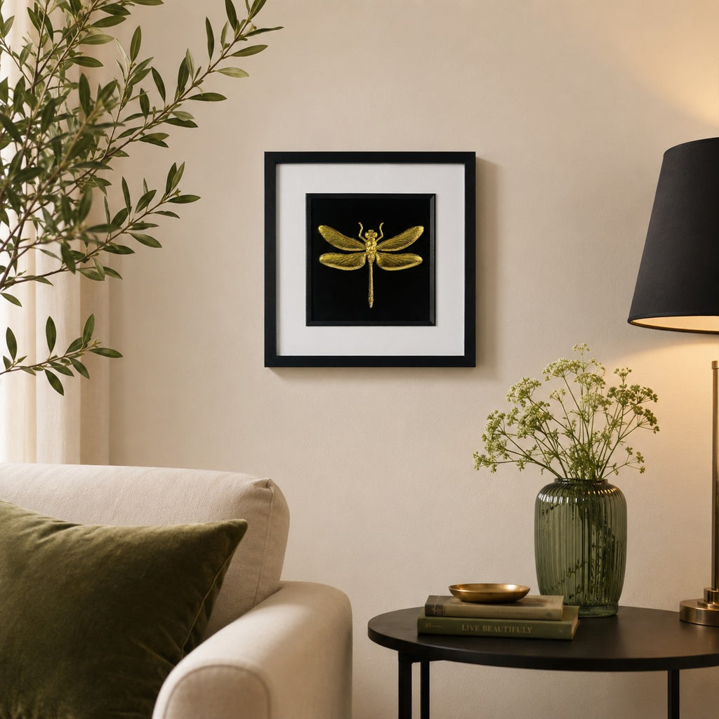 Framed gold dragonfly wall art in a black square frame with white mount, styled on a neutral wall in a calm modern living room with greenery and warm lighting.