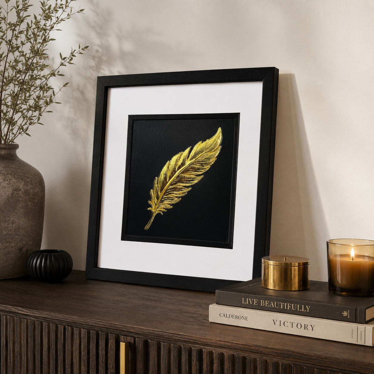 Angled lifestyle view of gold feather wall art in a black square frame with white mount, displayed on a dark wooden console with neutral decor, books and a lit candle.