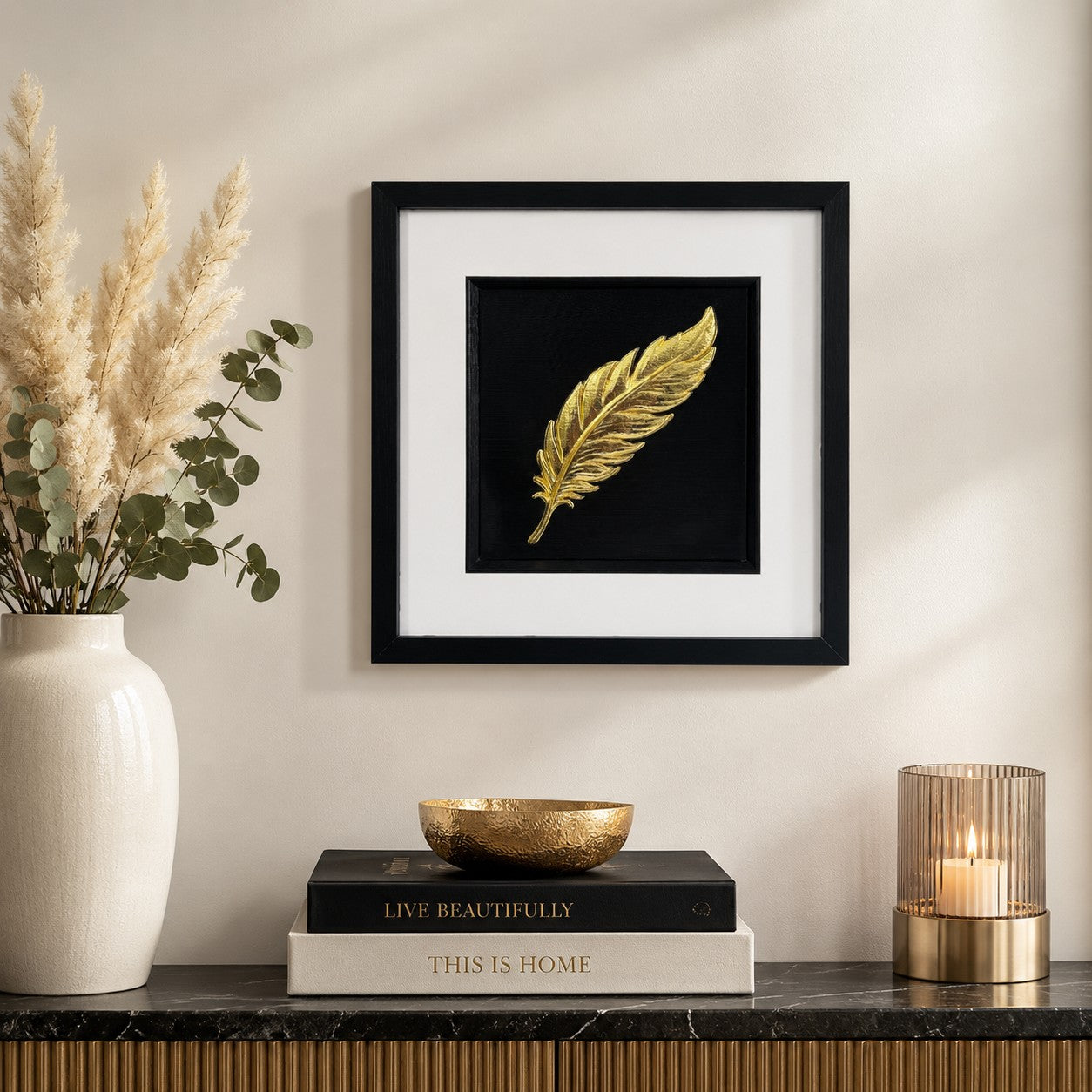 Gold feather wall art in a black frame with white mount, styled on a neutral wall above a dark console table with soft pampas stems, books and warm candlelight.