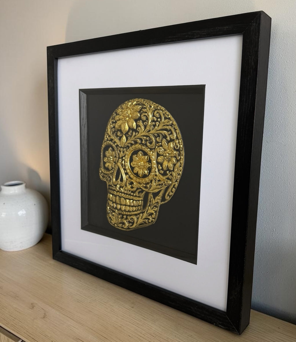 Gold floral sugar skull raised relief in a black box frame with white mount, styled on a wood sideboard with a white vase