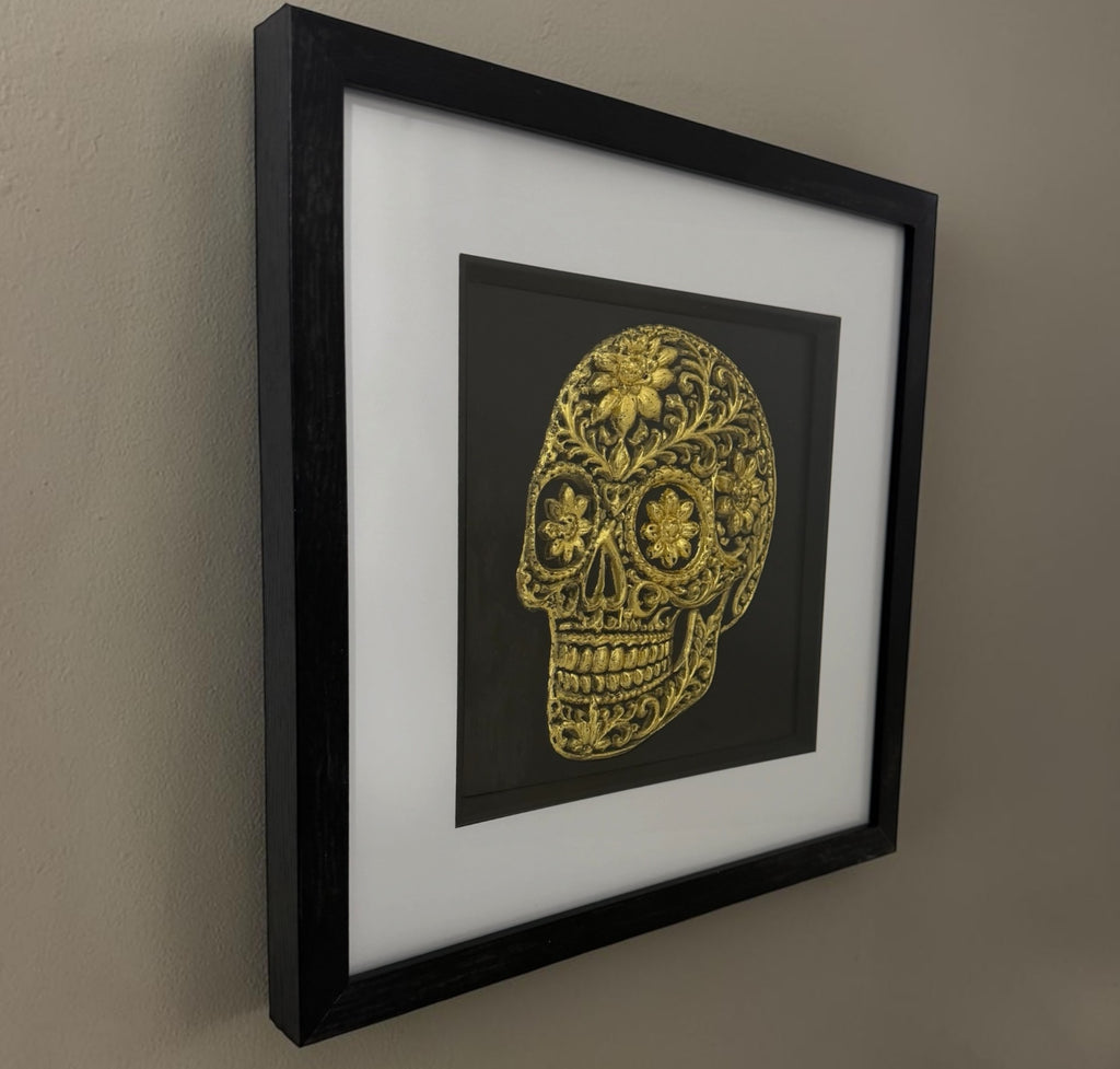 Gold floral sugar skull framed artwork hanging on a neutral wall, showing the black frame depth and white mount