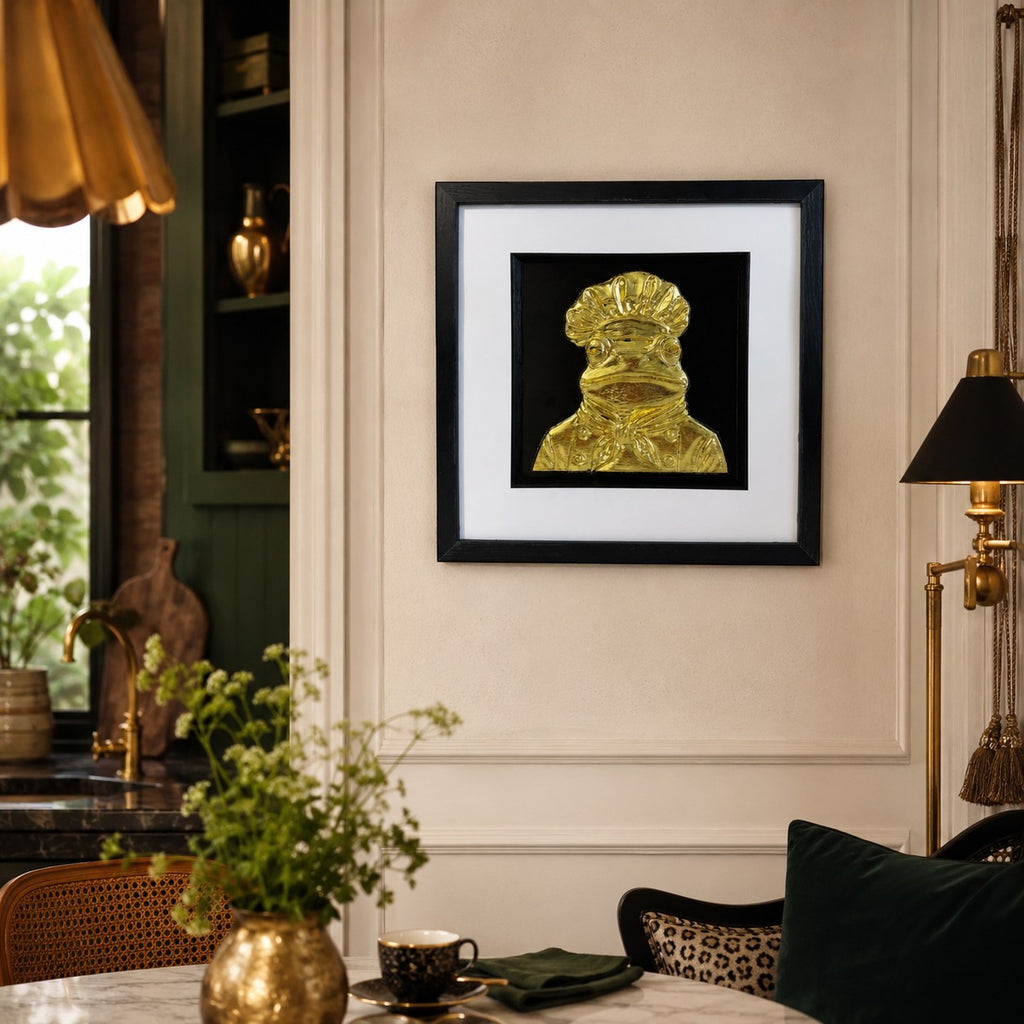 Gold frog chef framed kitchen wall art hanging in a warm kitchen dining scene with brass lighting and green accents