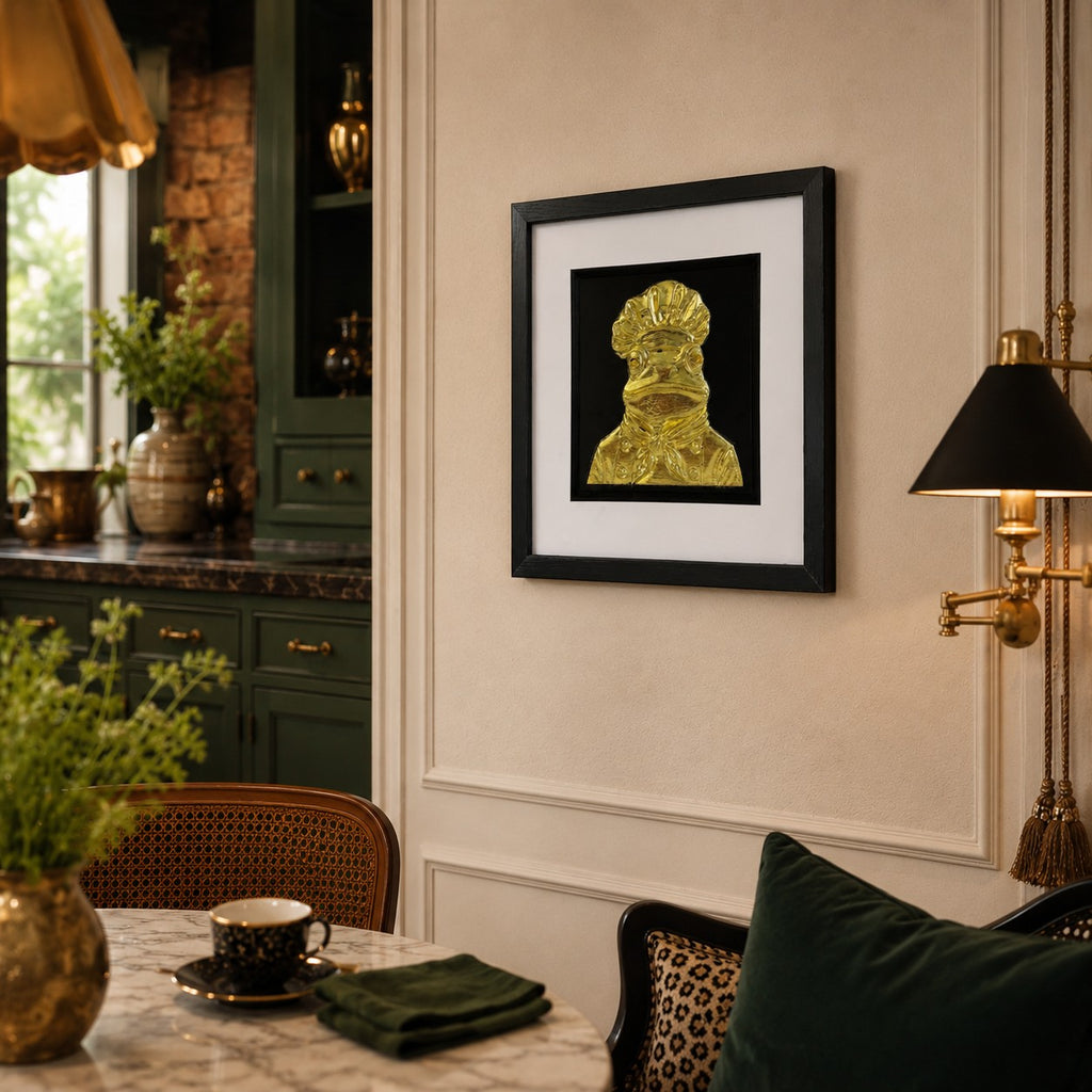 Angled lifestyle view of gold frog chef framed artwork on a kitchen wall beside dark green cabinetry and a brass lamp