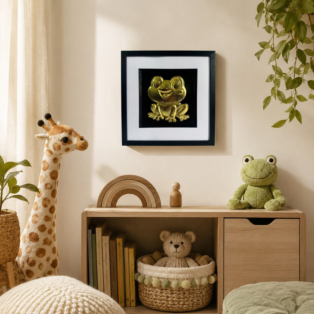Gold frog wall art displayed in a cosy nursery, featuring a 33 cm square framed hand gilded gold leaf 3D relief frog design on a black background with soft child-friendly decor.