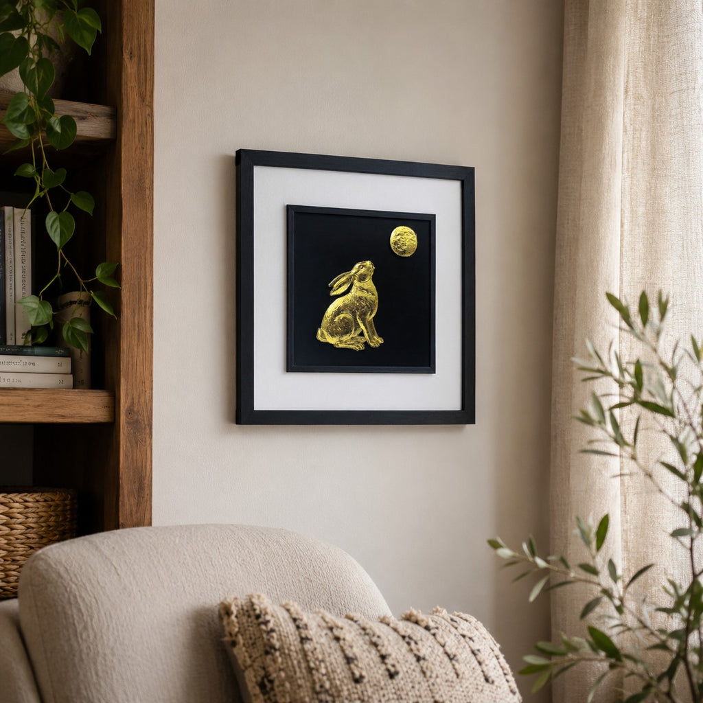 Angled lifestyle view of gold hare gazing at the moon wall art hung on a neutral wall beside natural wood shelving, plants and soft cosy decor.