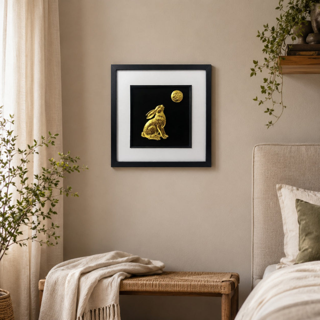 Gold hare and moon wall art in a black frame displayed on a warm neutral bedroom wall with soft textiles, natural wood and greenery.