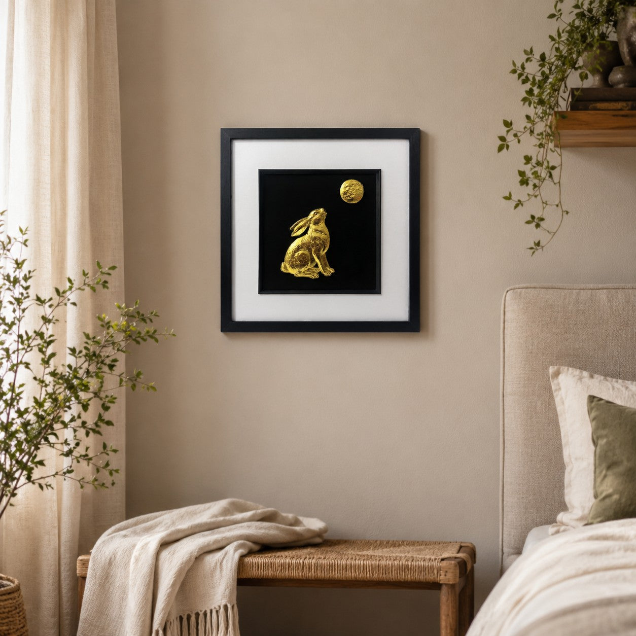 Gold hare and moon wall art in a black frame displayed on a warm neutral bedroom wall with soft textiles, natural wood and greenery.