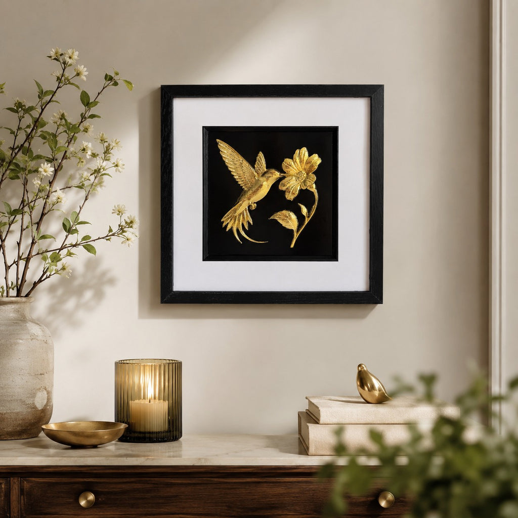 Framed gold hummingbird and flower wall art displayed on a warm neutral wall above a wooden console with botanical styling, candlelight and soft gold accents.