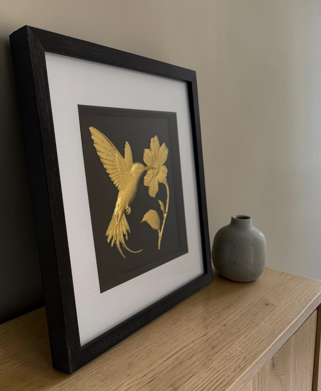 Framed gold hummingbird and flower wall art displayed at an angle on a light wooden sideboard with a neutral vase, styled in a calm minimalist home interior.
