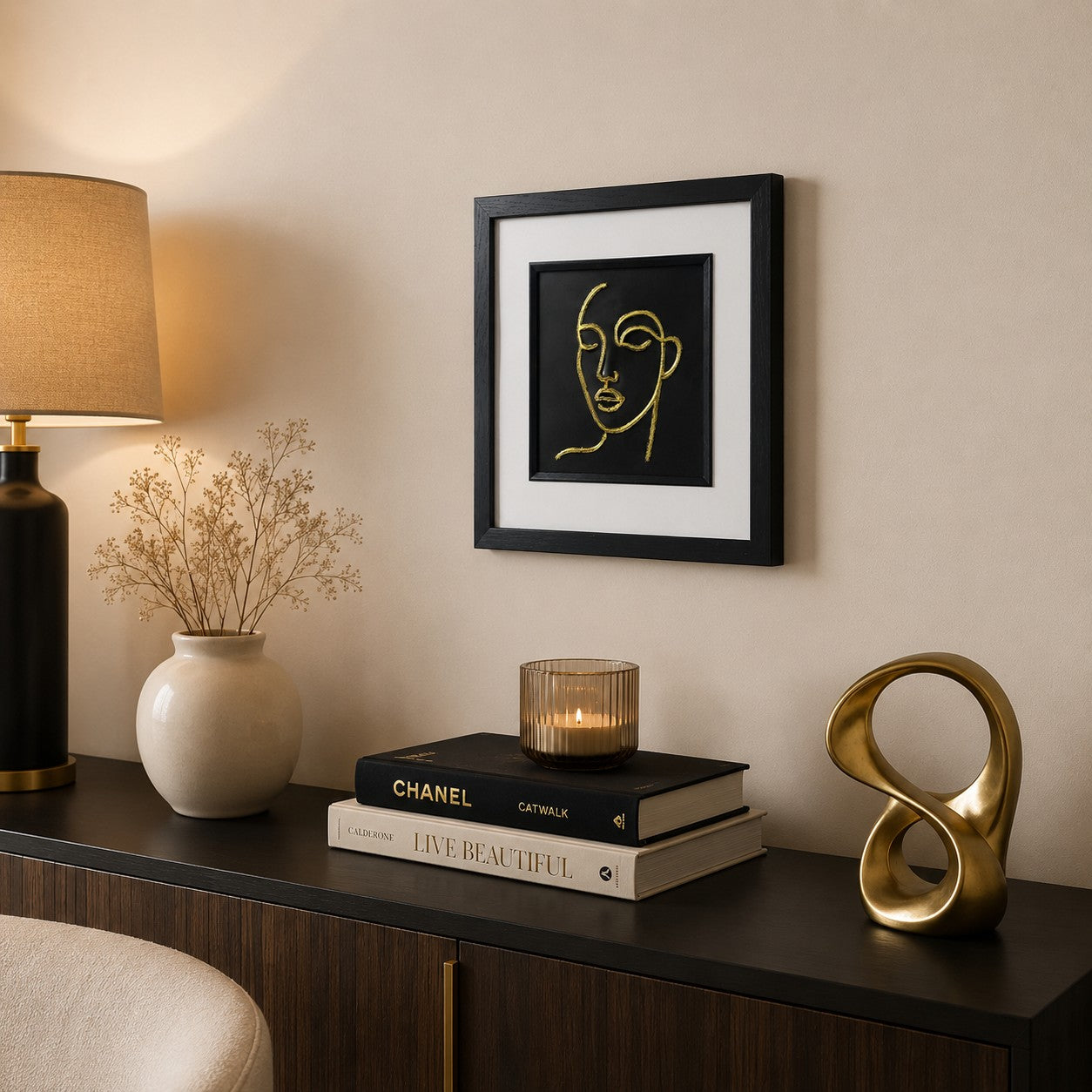 Angled lifestyle view of gold line face wall art in a black square frame with white mount, displayed in a modern neutral interior with books, candlelight and gold decor accents.
