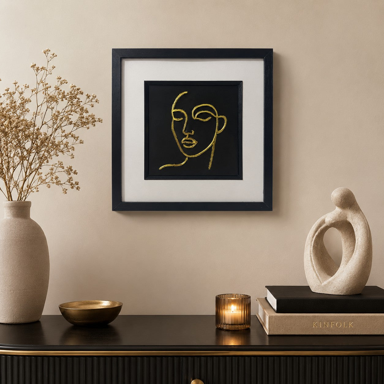 Gold line face wall art in a black frame with white mount, styled on a neutral wall above a dark console with a ceramic vase, candle and sculptural decor.