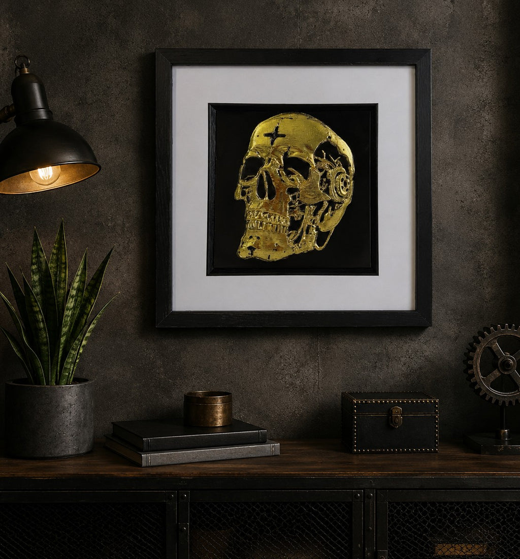 Framed gold mechanical skull wall art displayed on a dark industrial wall above a metal console, styled with a plant, vintage lamp and steampunk décor accents.