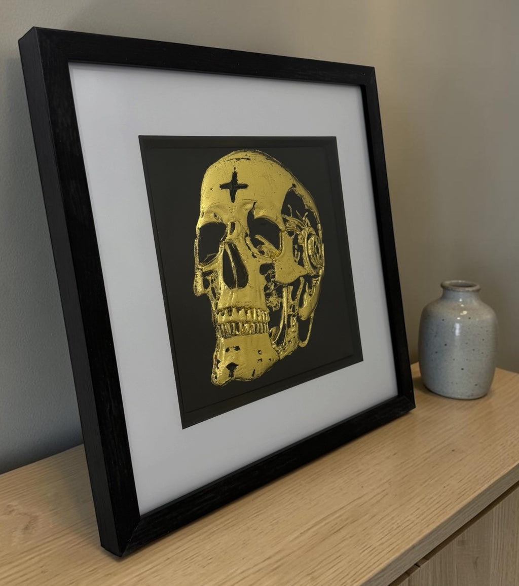 Framed gold mechanical skull wall art displayed at an angle on a light wooden sideboard with a neutral ceramic vase, styled in a modern home interior.